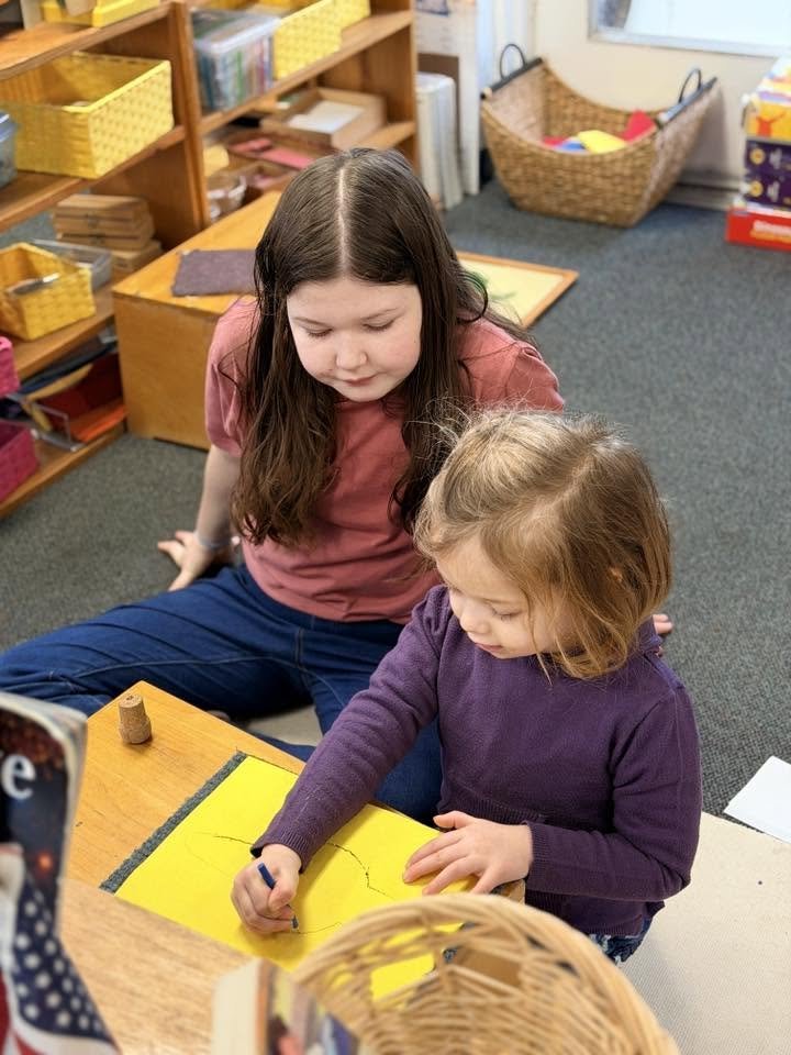 Two young girls sitting at a desk in a classroom, one assisting the other with drawing on a yellow paper.