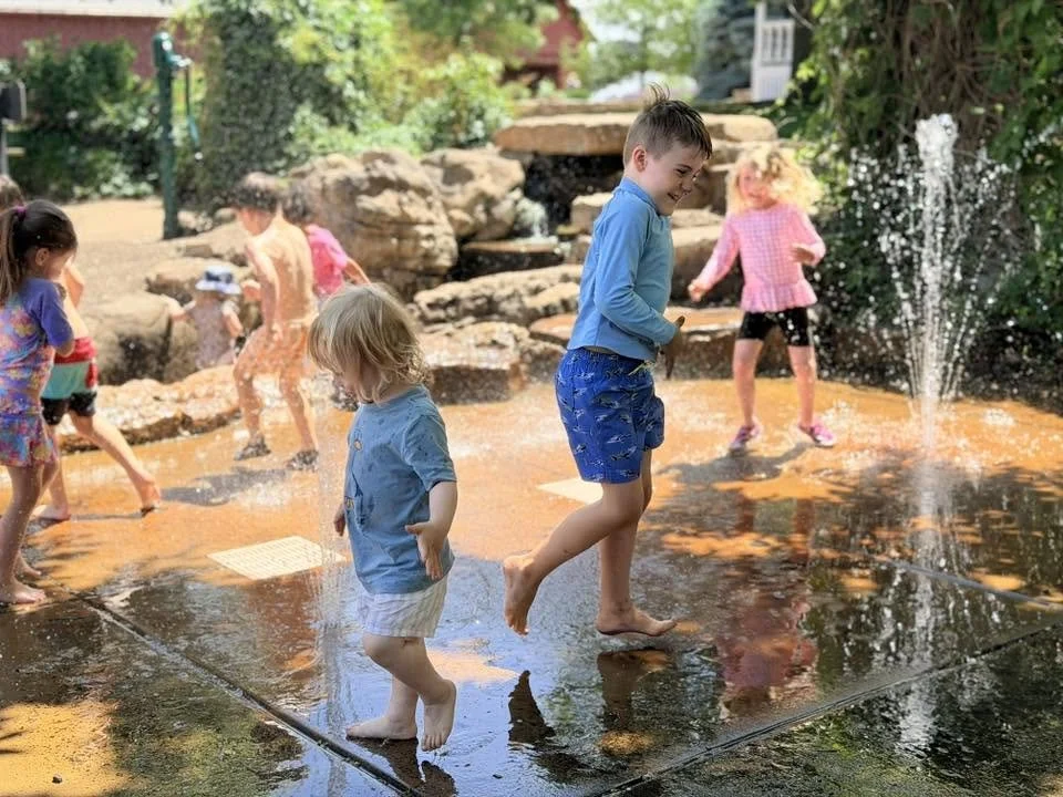 Children playing in a water park with splashing fountains, rocks, and greenery in the background.