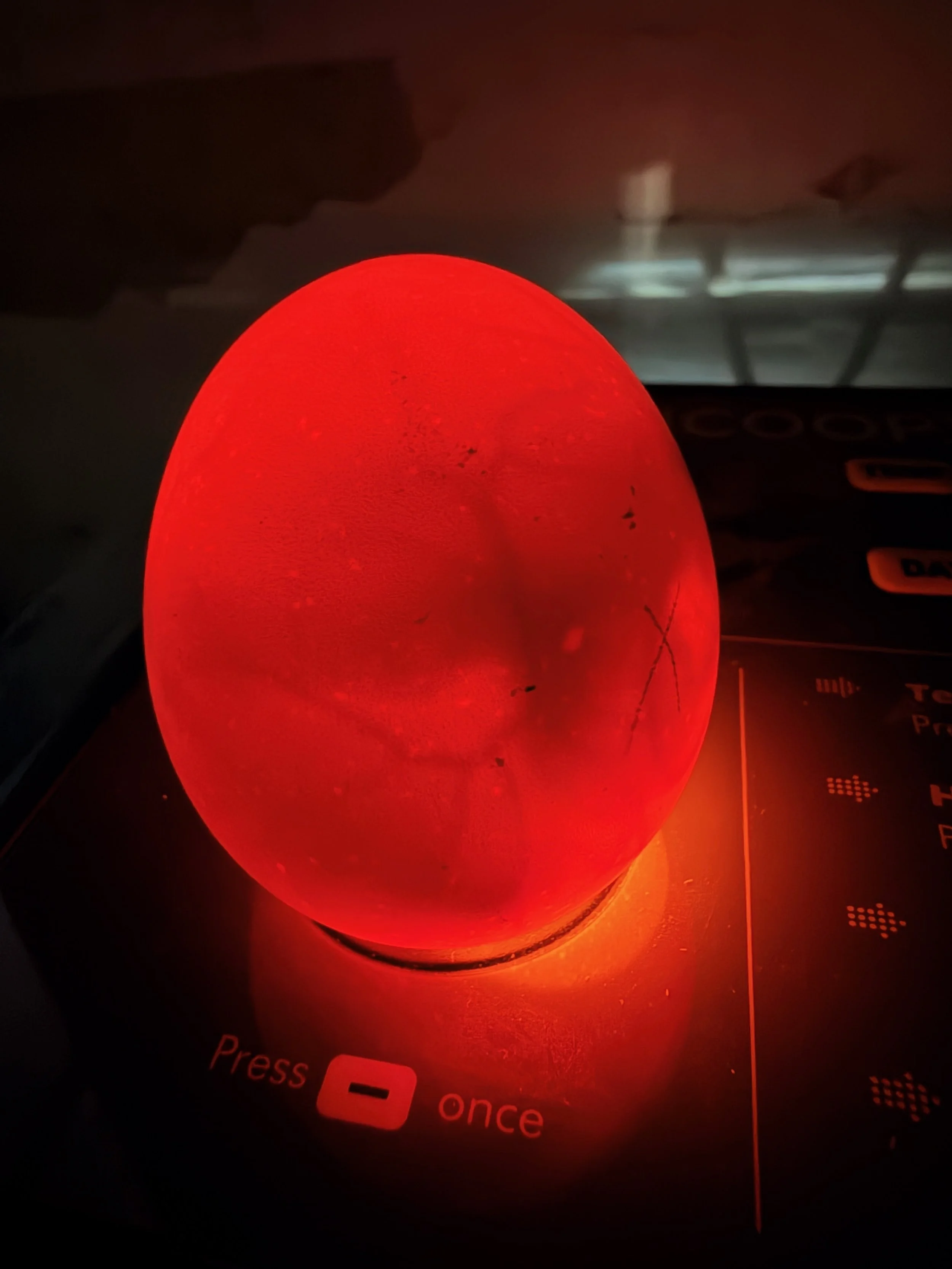 Red glowing egg-shaped object with black marks resting on a surface, with a dark background featuring some electronic device buttons and a grid-patterned area.