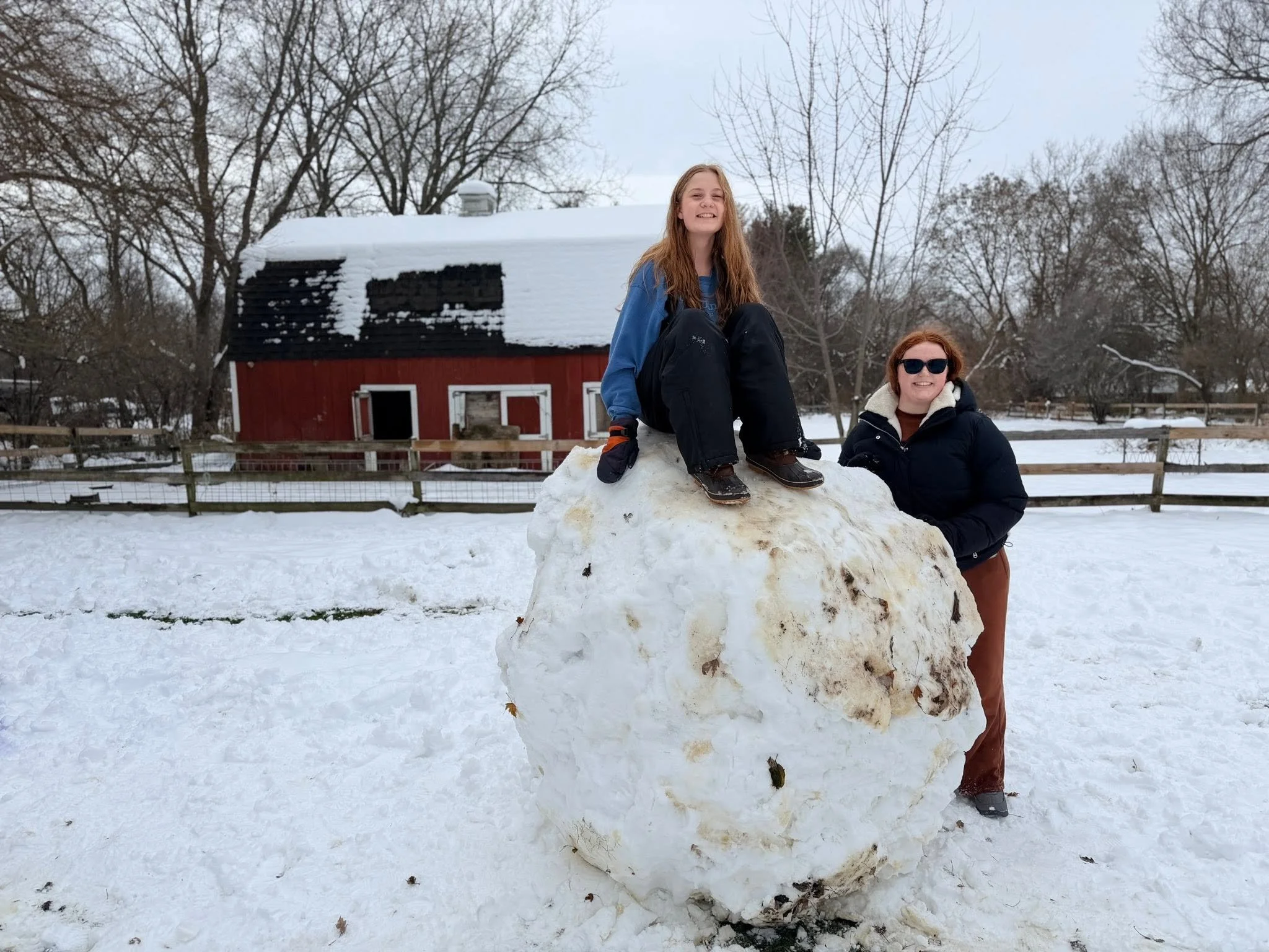 Two smiling women in winter clothing are posing with a large snowball in a snowy outdoor setting. One woman is sitting on top of the snowball, and the other is standing beside it, leaning on it. There is a red barn and leafless trees in the backgroun