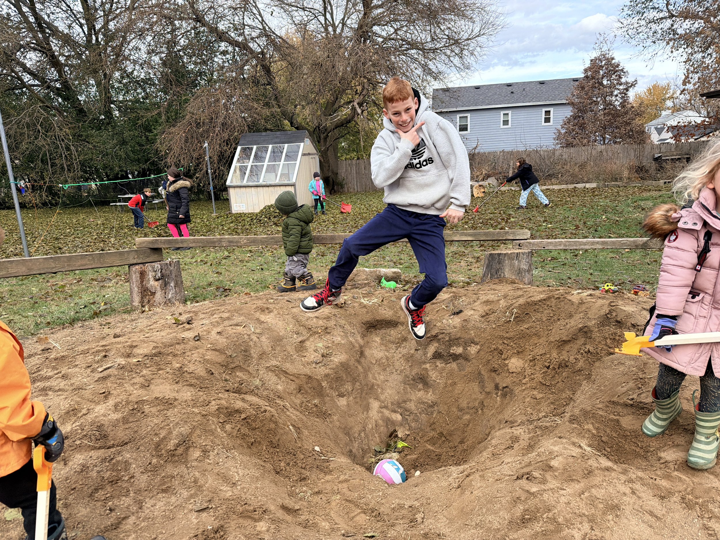 Kids playing in a backyard with a sandbox and toys, some digging in the sand, others walking or standing, with trees, a house, and a shed in the background.