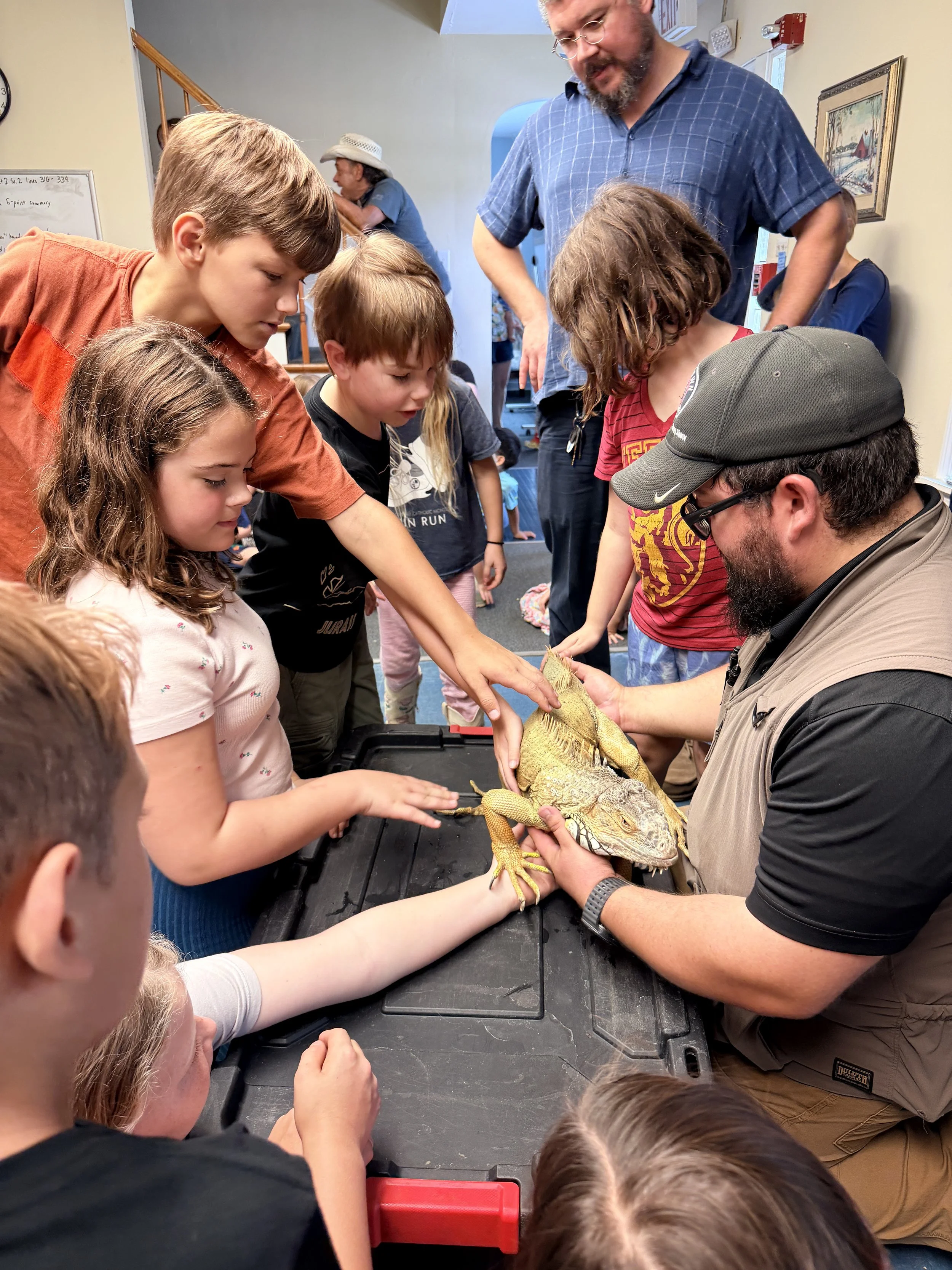 Group of children and adults gathered around a table, petting and examining a large yellow and gray iguana.