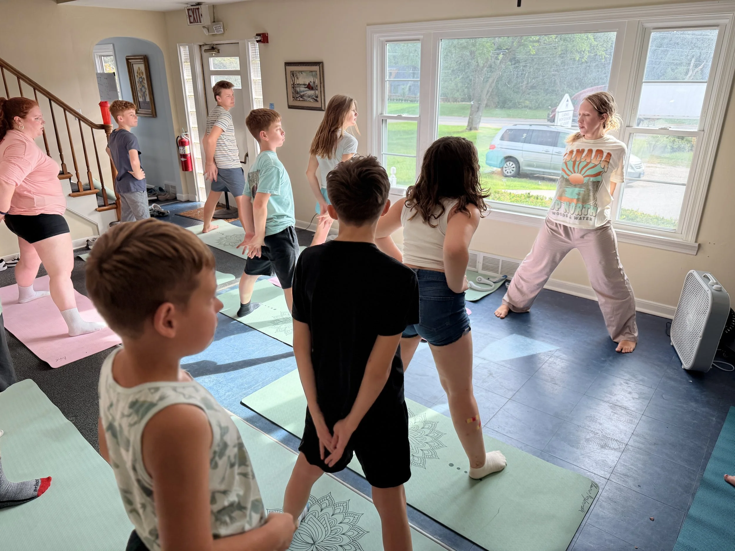 A group of children participating in a yoga or exercise class indoors, with some on yoga mats and one leading the session near a large window showing outdoor greenery.