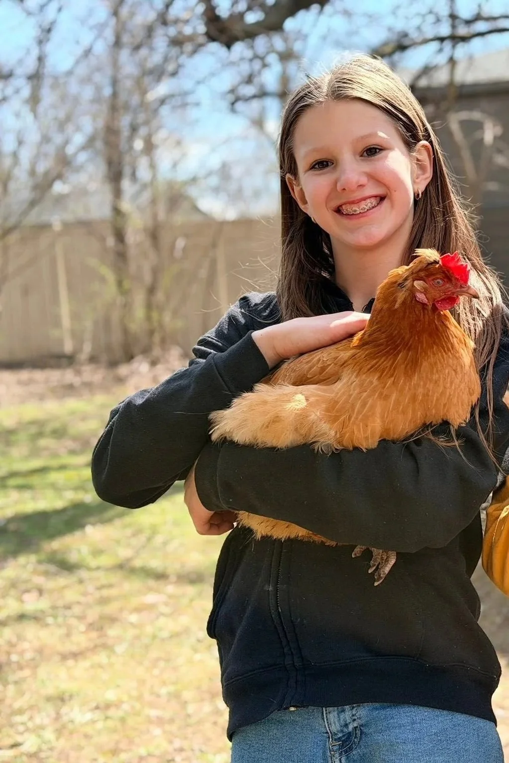 A girl with long brown hair and braces is smiling while holding a large brown chicken outdoors in a backyard with trees and a wooden fence.