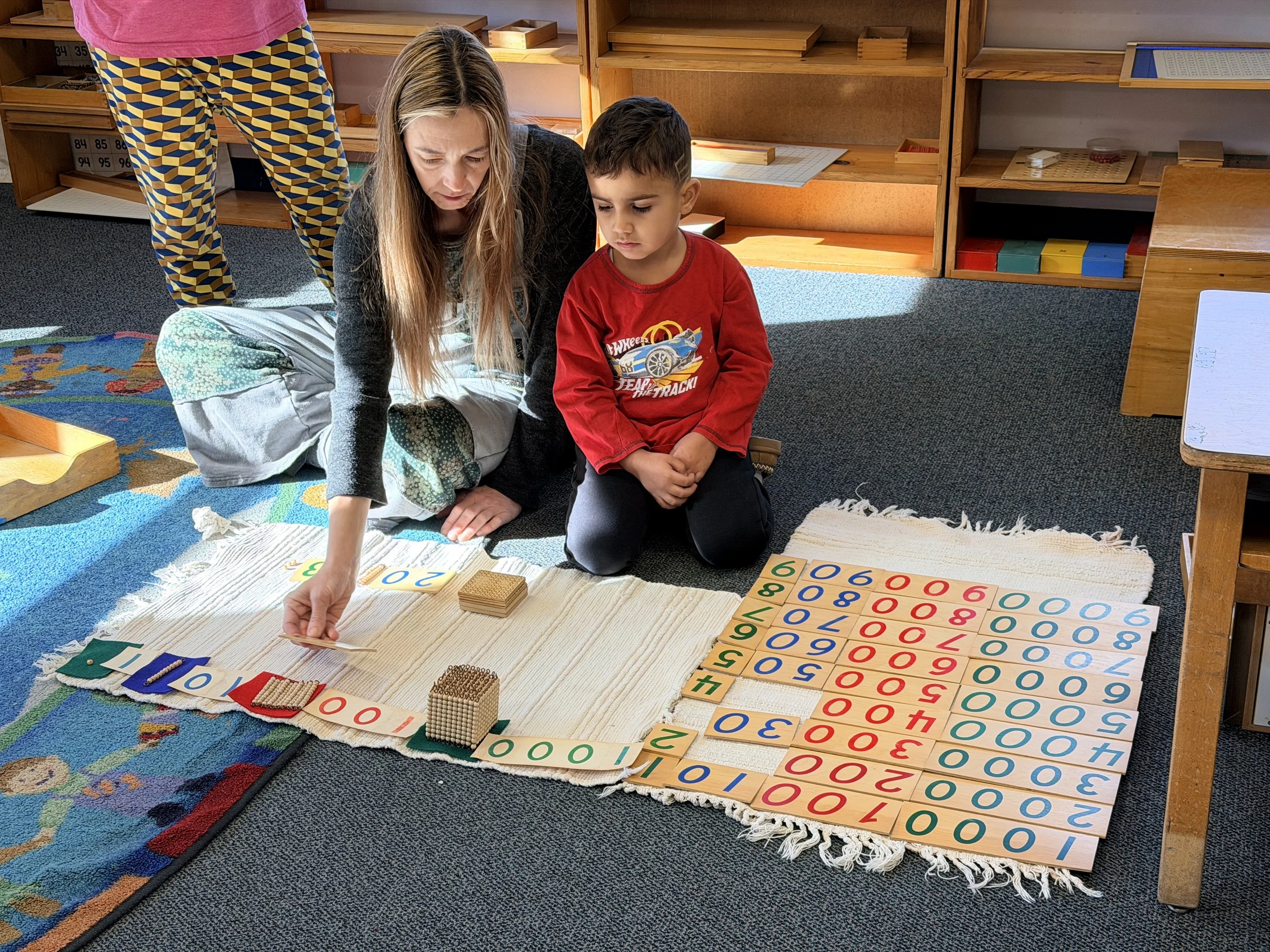 A teacher and young boy working on a math activity with number tiles, blocks, and a number line in a classroom.