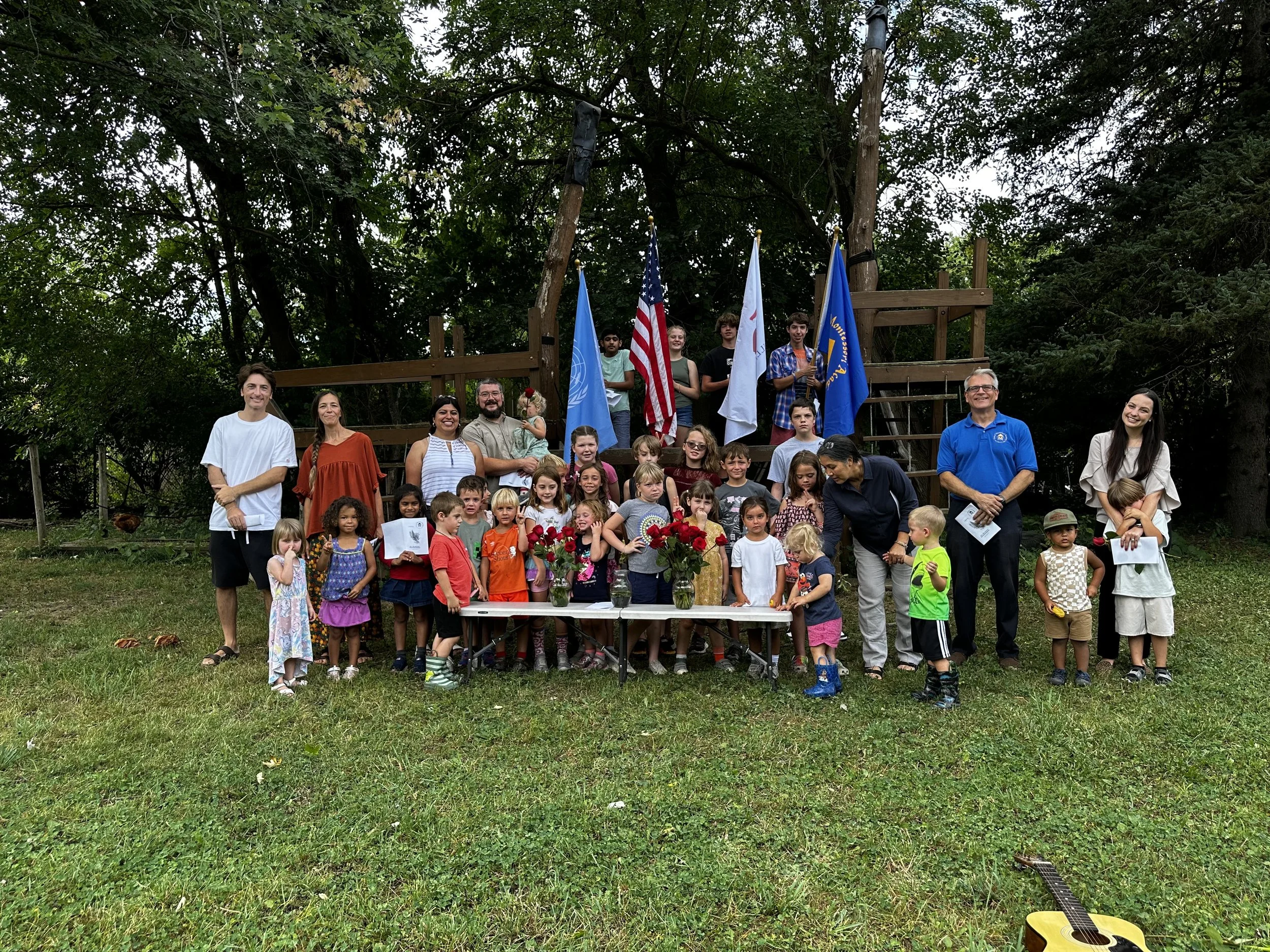 A large group of children and adults gathered outdoors under trees, with some holding flags and bouquets of flowers, celebrating on a grassy area.