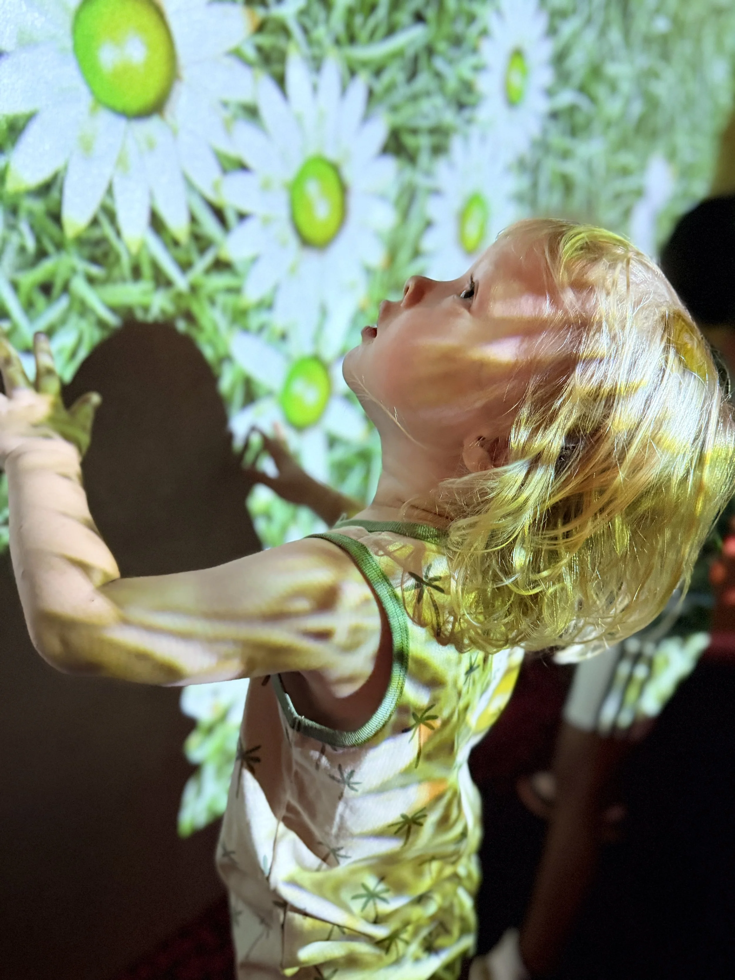 A child with curly blonde hair looking up, wearing a light-colored dress with green star patterns, in front of a projected image of white daisies with green centers.