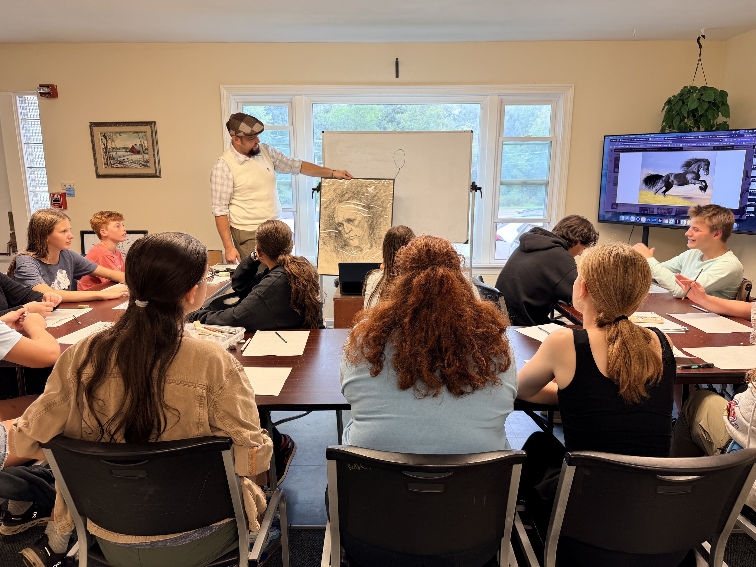 A classroom filled with students seated at tables, listening to a male instructor standing near a whiteboard with a sketched outline of a face. A large screen displays an image of a black horse running.