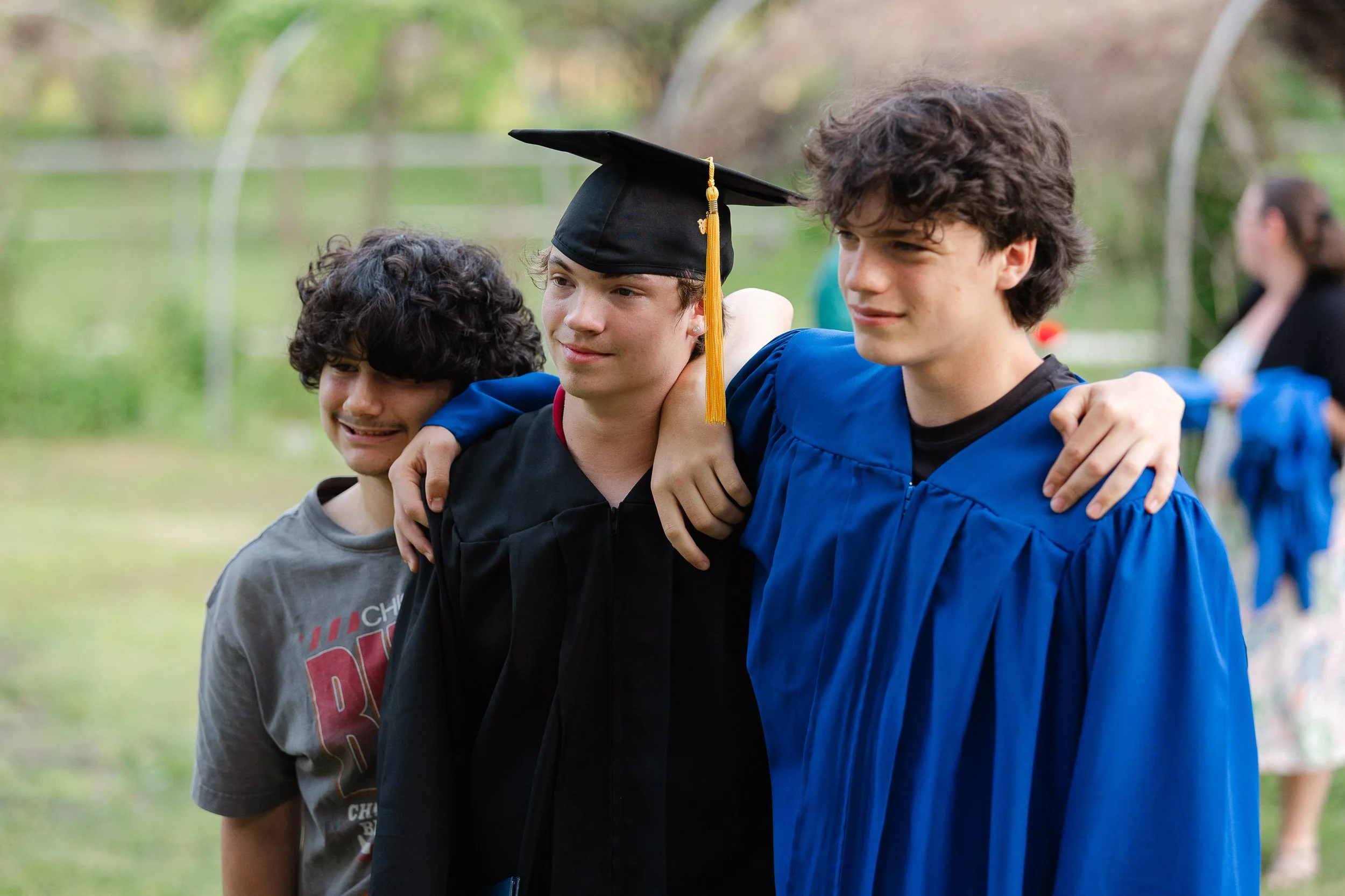 Three young men standing outdoors with their arms around each other, one wearing a graduation cap and gown, smiling.