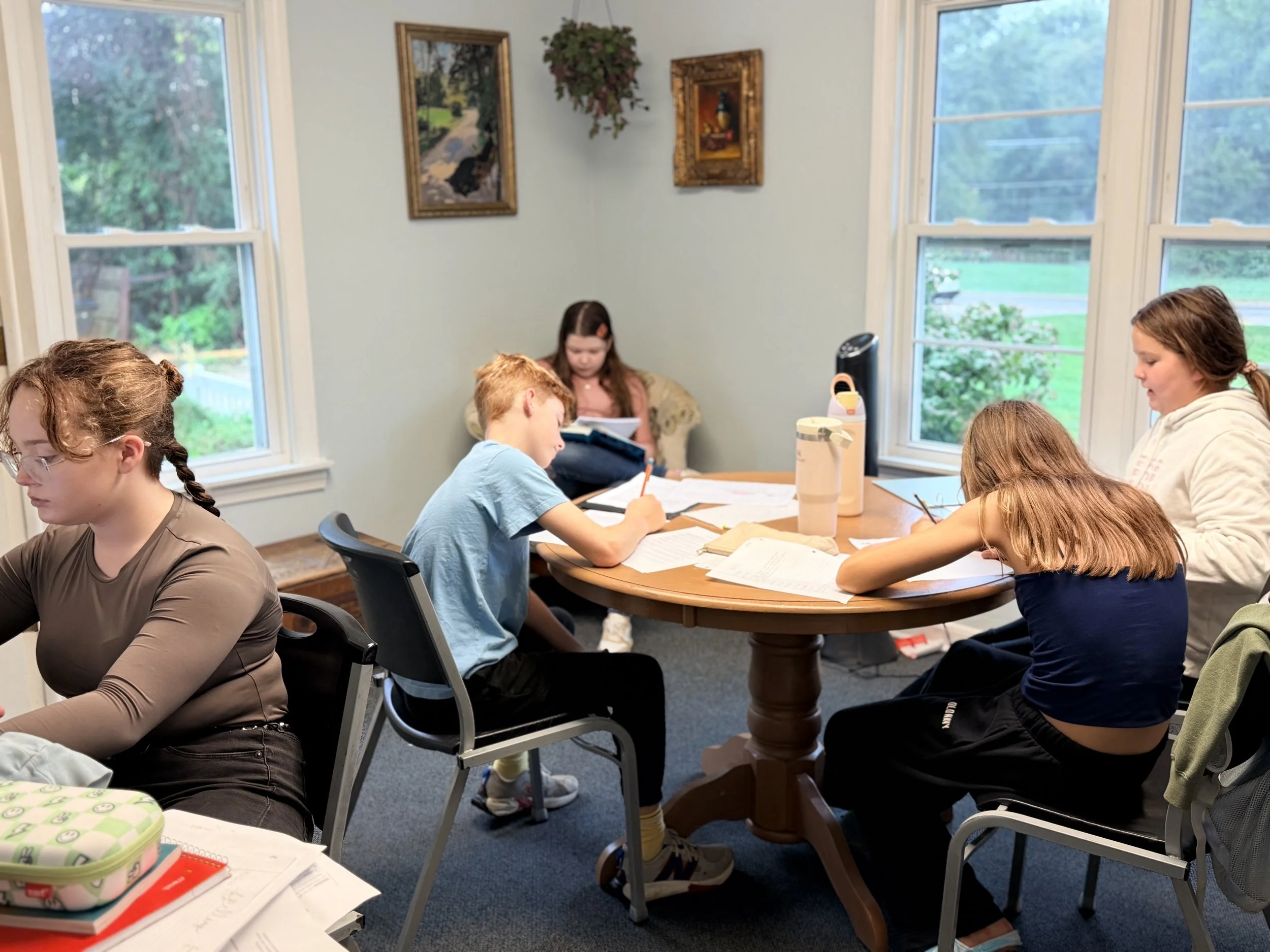 Group of children and a young woman studying at a wooden table in a bright room with three large windows showing greenery outside. The children are engaged with papers and books, and the woman is reading. Items on the table include notebooks, a water