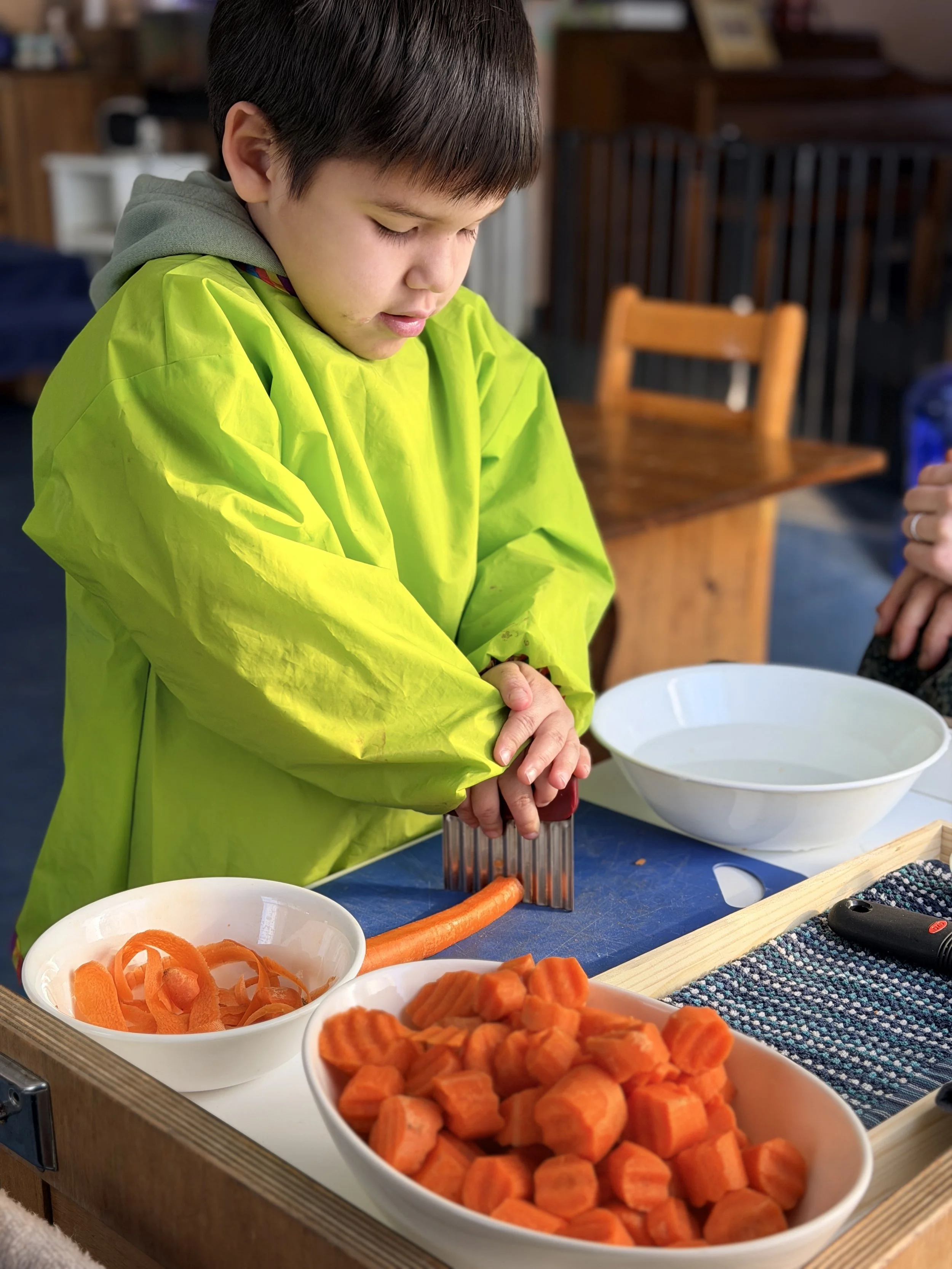 A young boy wearing a yellow raincoat and gray hoodie grating a carrot on a grater beside bowls of sliced carrots and carrots chunks on a kitchen counter.