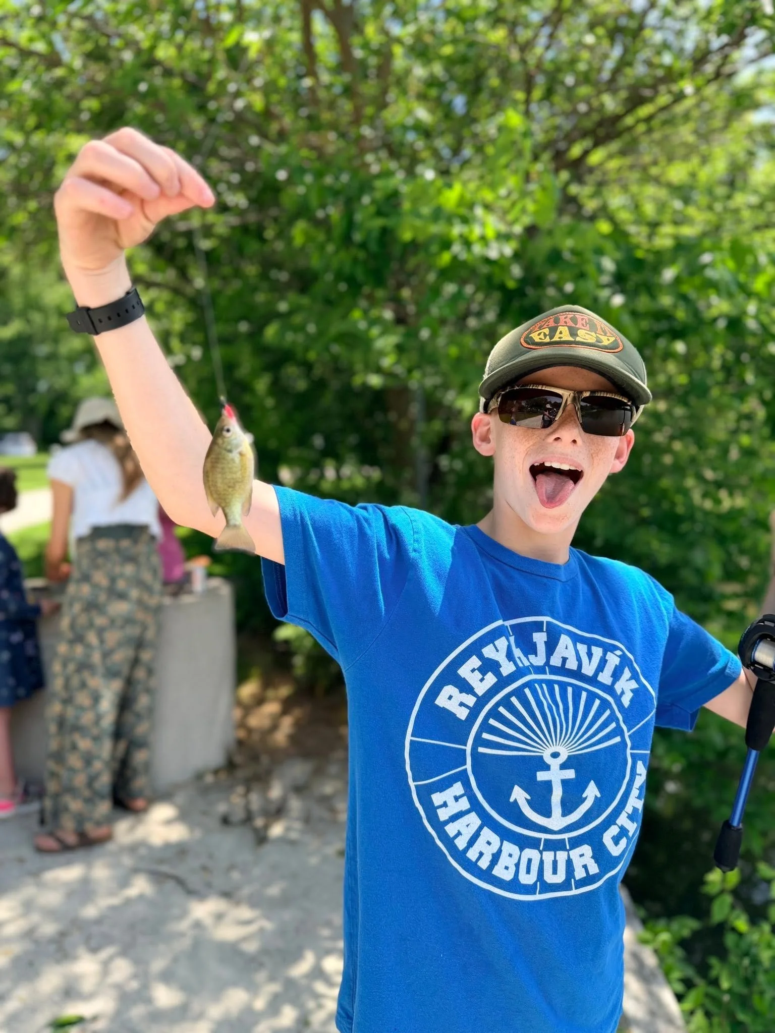 A boy wearing sunglasses and a cap with a fishing logo holding a small fish outdoors during daytime, with green trees in the background.