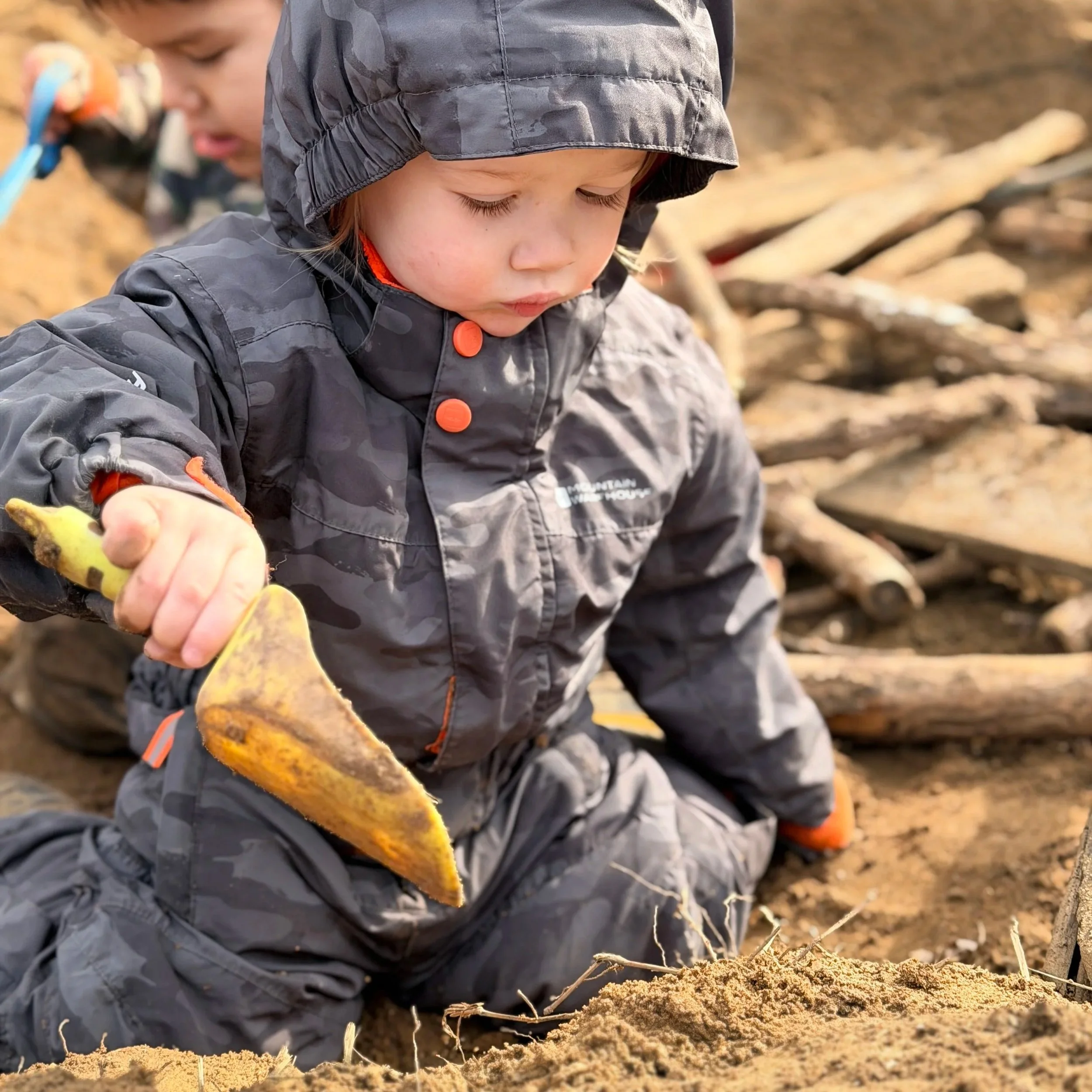 A young child in a gray hooded jacket with orange buttons is sitting in the dirt, digging or playing with the soil and yellow gloves. The child appears to be focused on their activity, and a second child can be seen blurred in the background.