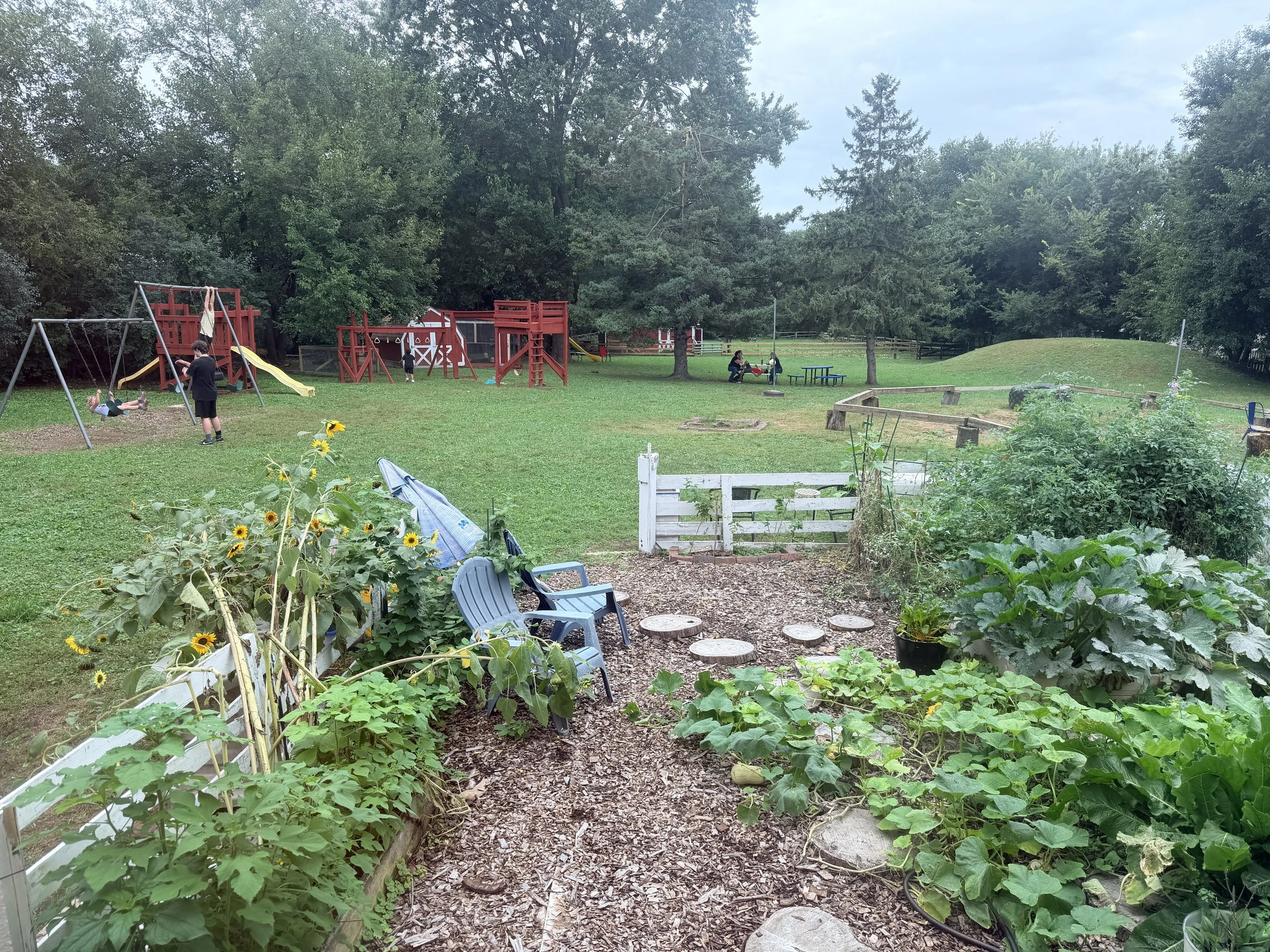 A children's playground with swings, slides, and play structures, surrounded by green grass and trees. There are children playing and adults sitting at picnic tables. The scene is set in a park-like area with a clear, overcast sky.