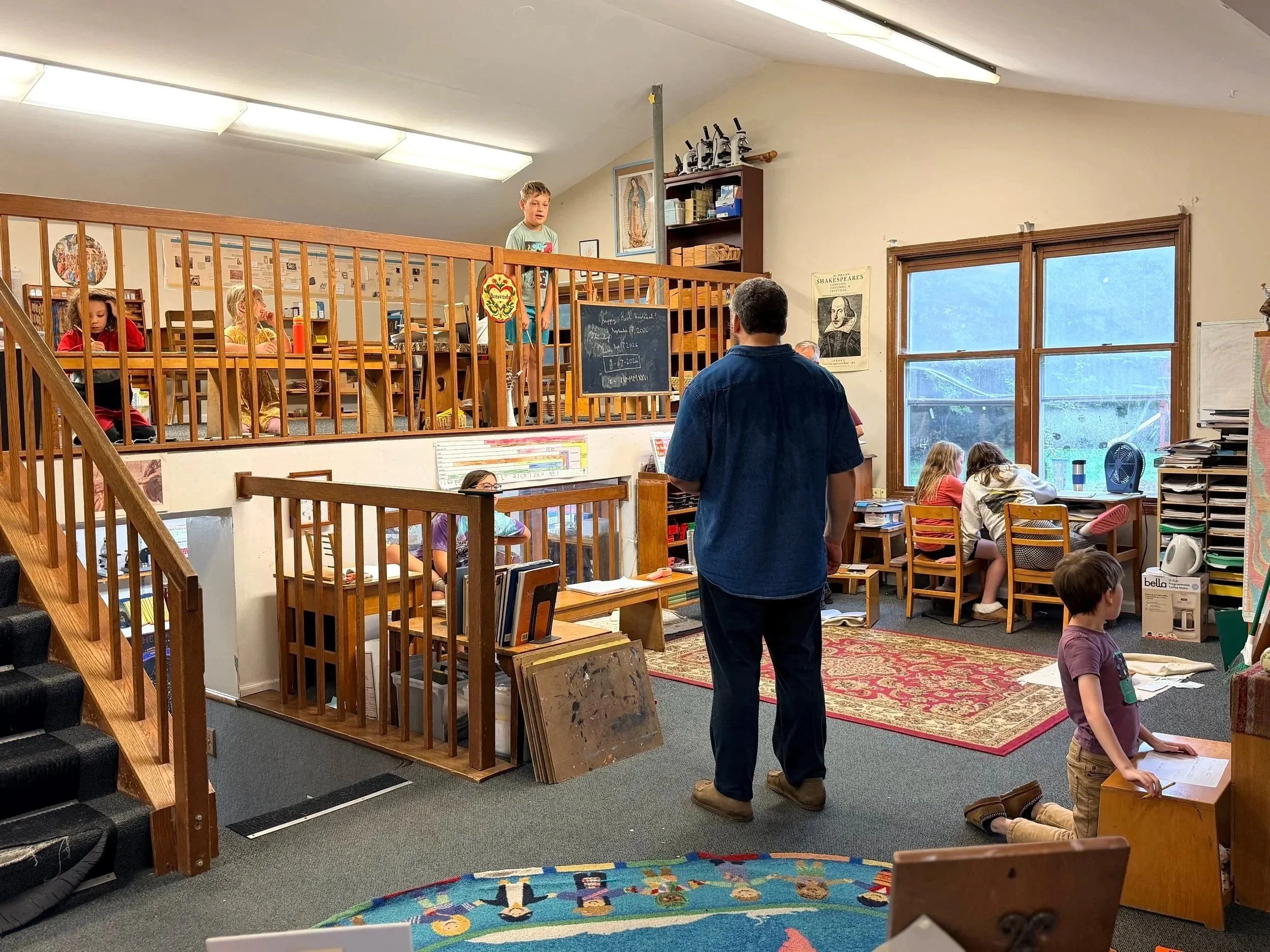 Inside a classroom with children working at desks and a teacher standing near a chalkboard, with a raised platform and wooden railing.