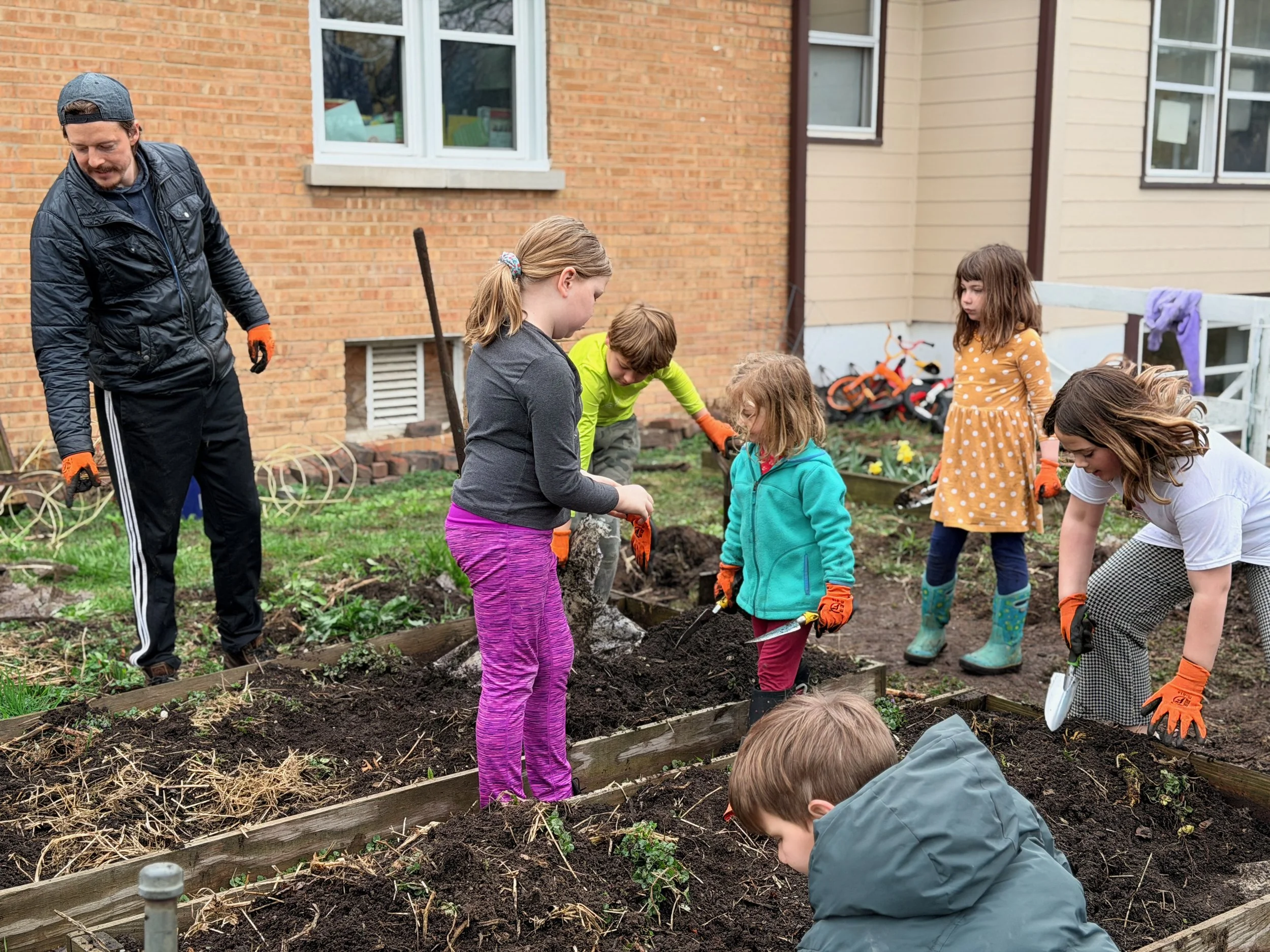 A group of children and adults planting in a garden bed outside a brick house.