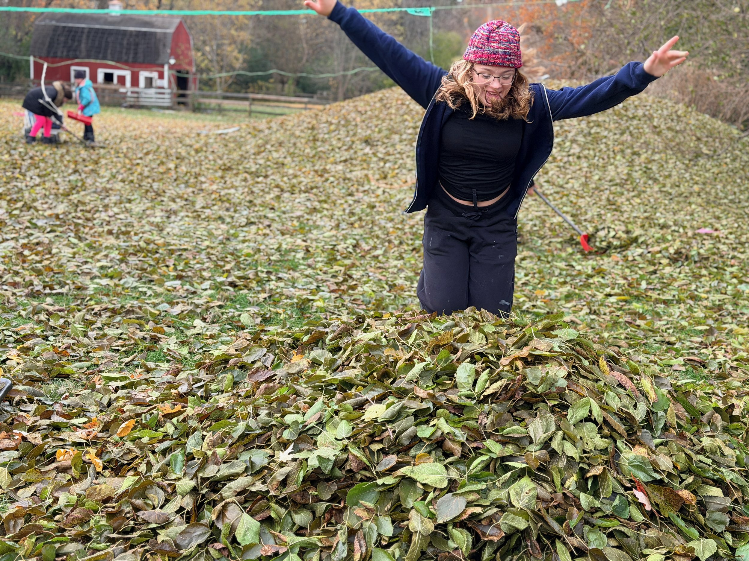 A woman with glasses, a red knit hat, black shirt, and navy jacket poses with her arms outstretched in a yard covered with fallen leaves. In the background, two children are raking leaves near a red barn.