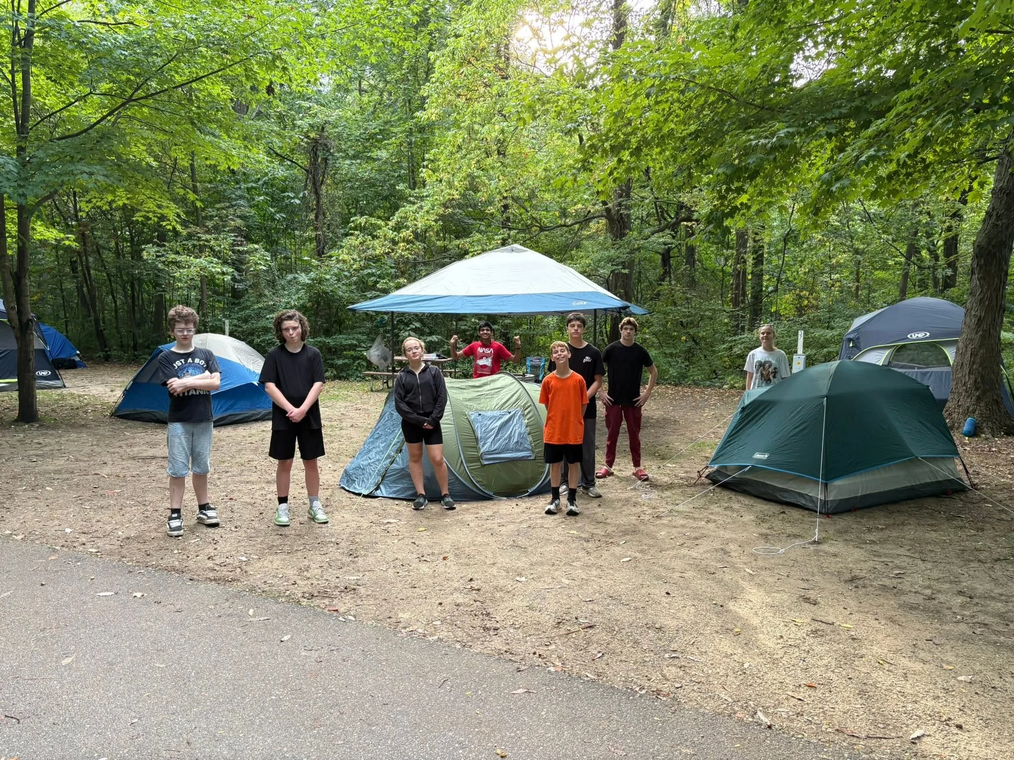 A group of young people standing in front of tents and a canopy in a wooded campsite during daytime.