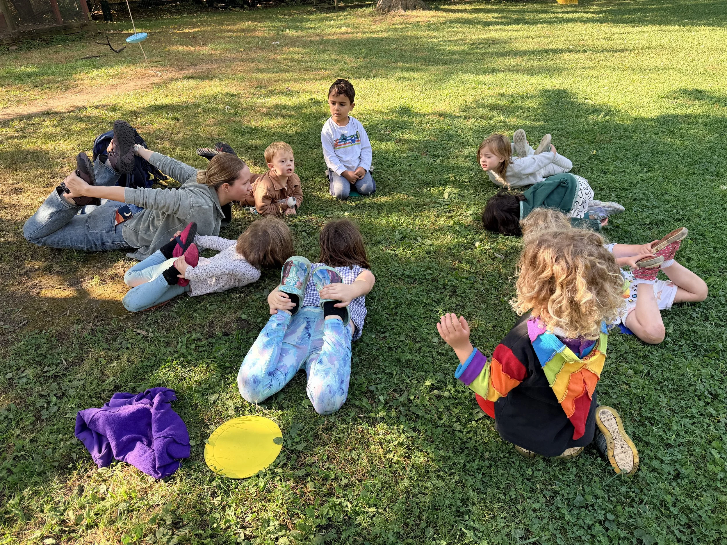 Children and an adult sitting and lying on grass in a park, engaging in a storytelling or group activity during daytime. The scene includes a tree-lined background and scattered yellow and purple items on the ground.