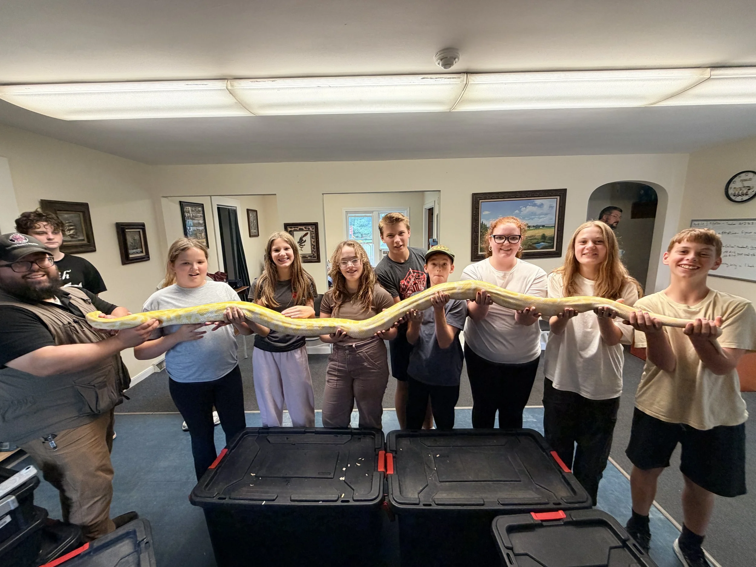 Group of children and adults gathered indoors, holding a large yellow and white snake, smiling at the camera. The room has framed pictures on the wall and a black storage container on the floor.