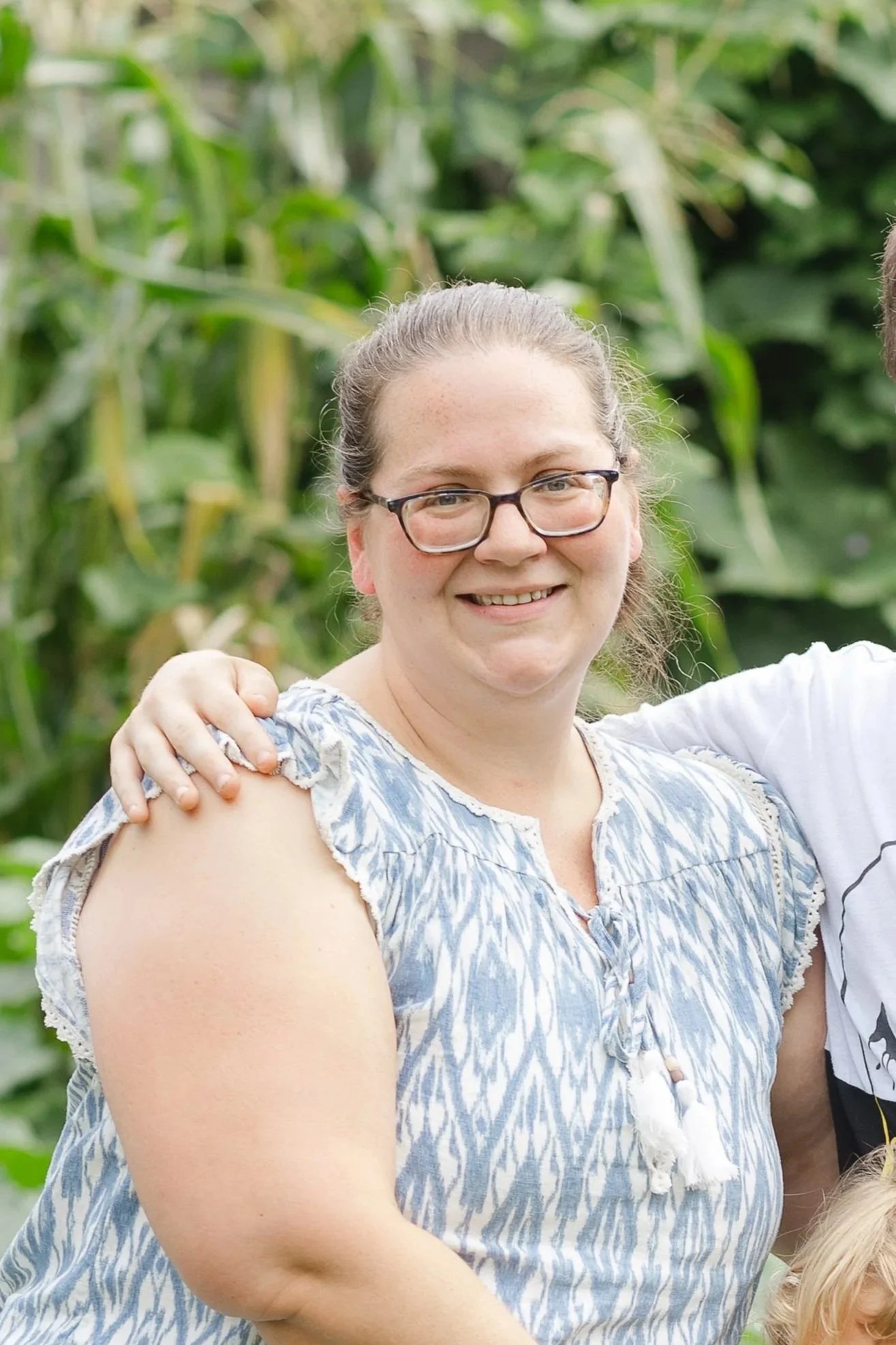 A woman with glasses smiling, partially hugging someone, standing outdoors among green plants and tall corn stalks.