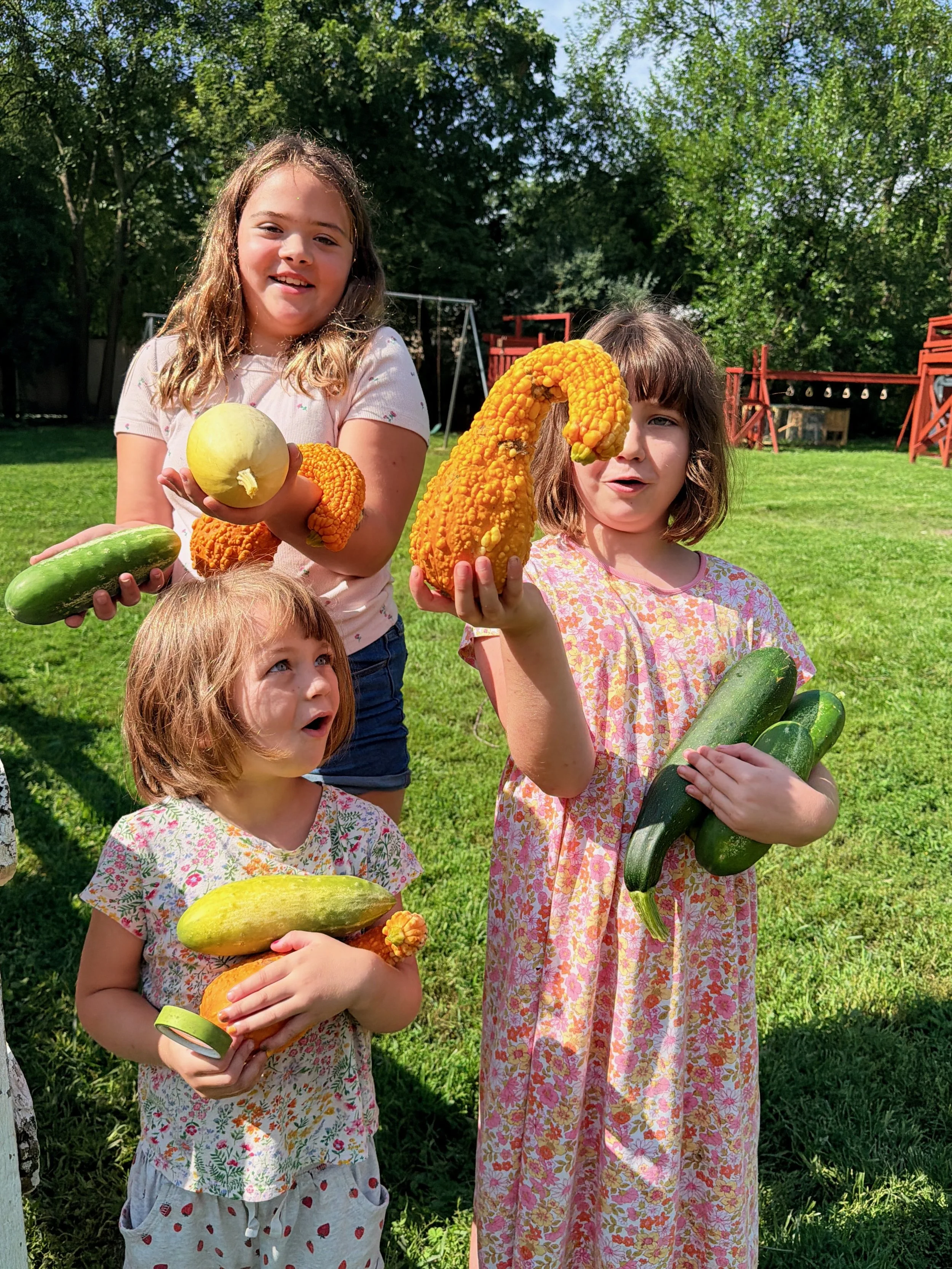 Four children holding various gourds and zucchinis in a backyard with green grass and trees, sunny weather.