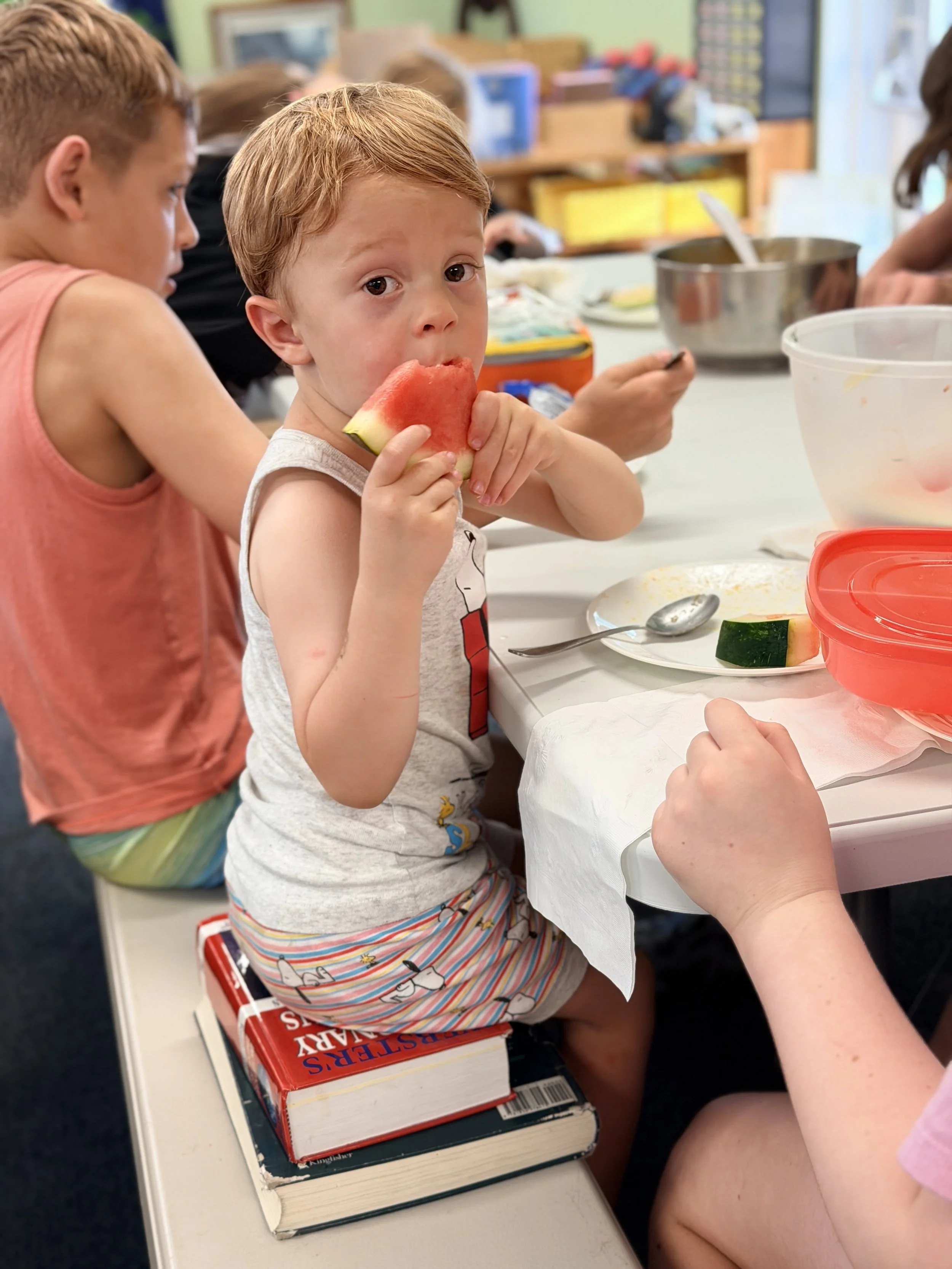 A group of children sitting at a table eating watermelon, with books and kitchenware around them.