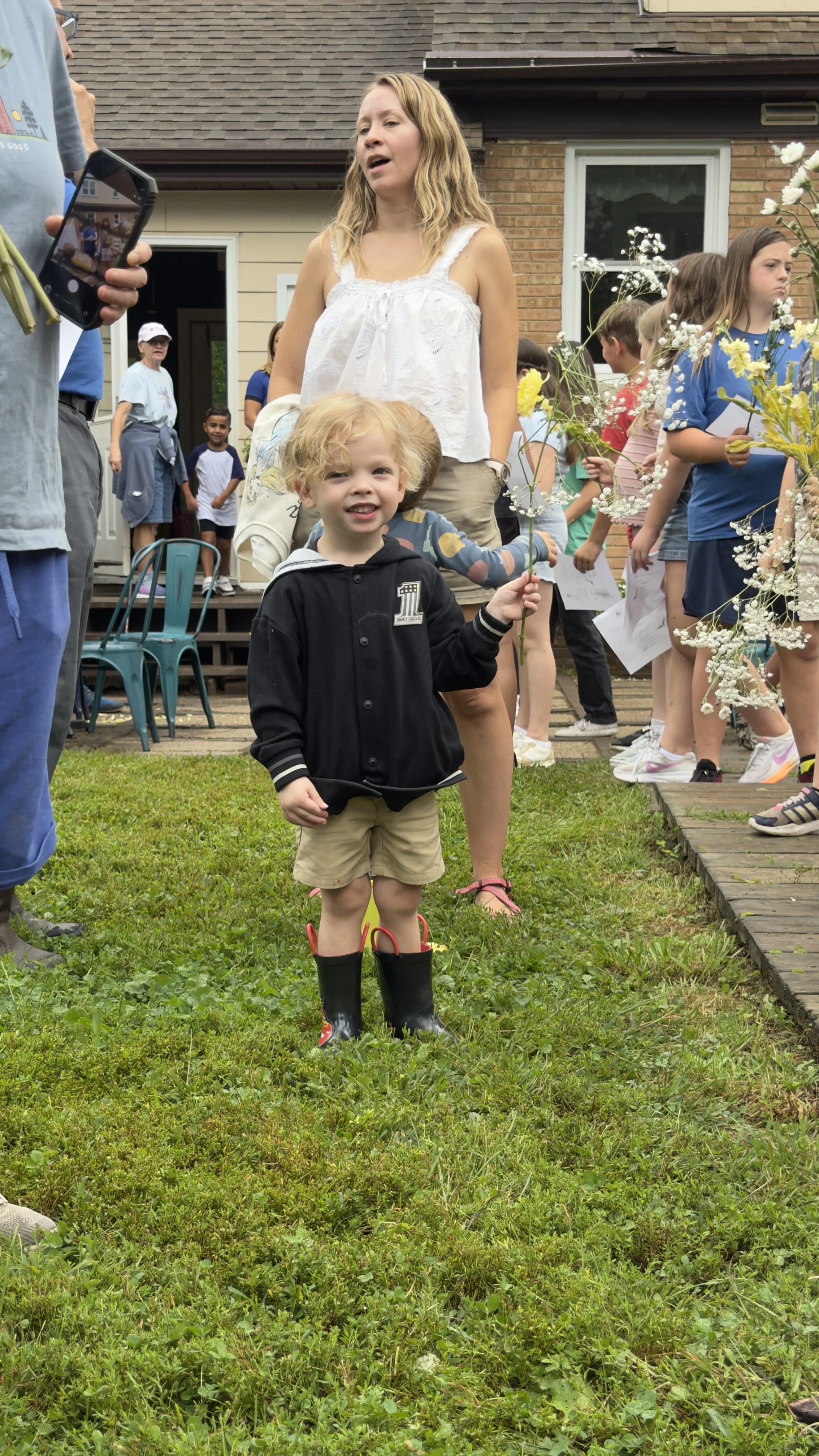 A young boy wearing rain boots, khaki shorts, and a black hoodie standing on grass, holding a flower, at an outdoor gathering with children and adults in the background.