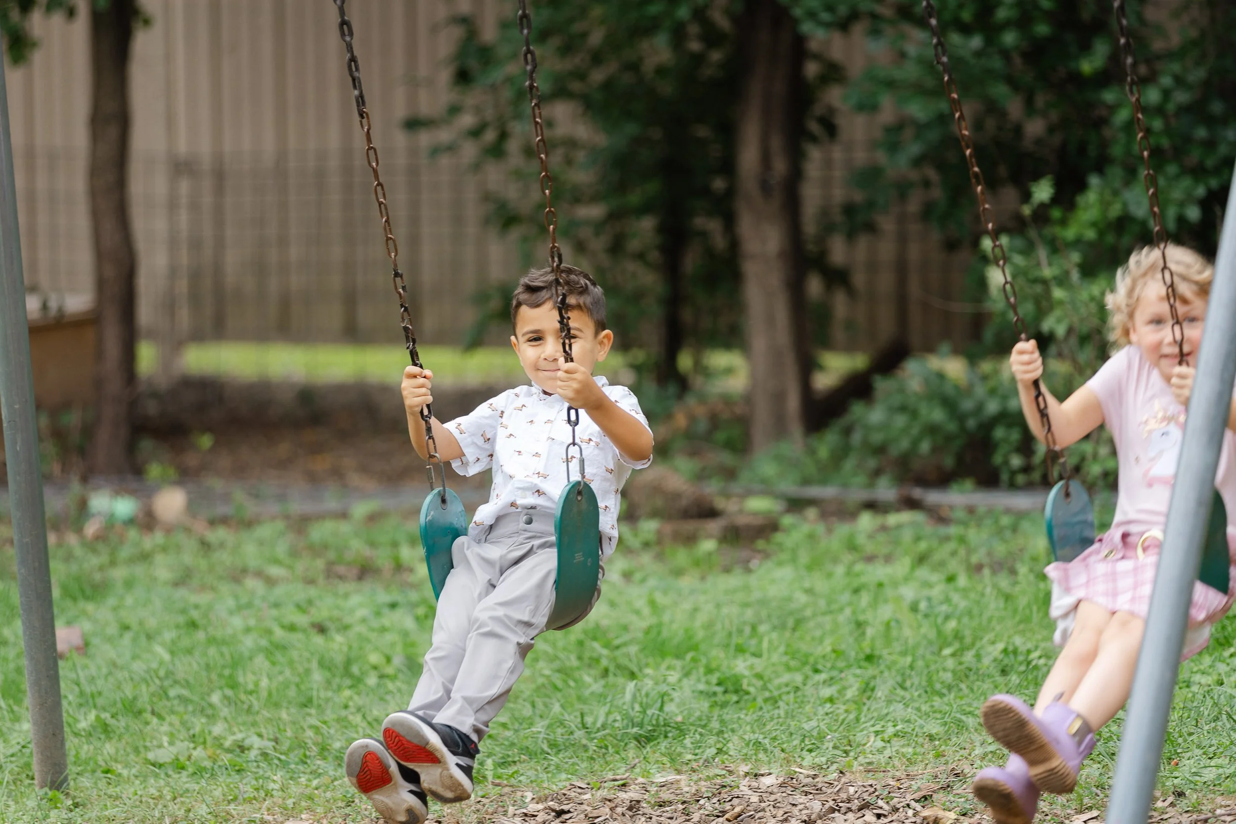 A young boy with dark hair smiling and sitting on a green swing, holding the chains, in a backyard with grass, trees, and a wooden fence in the background.