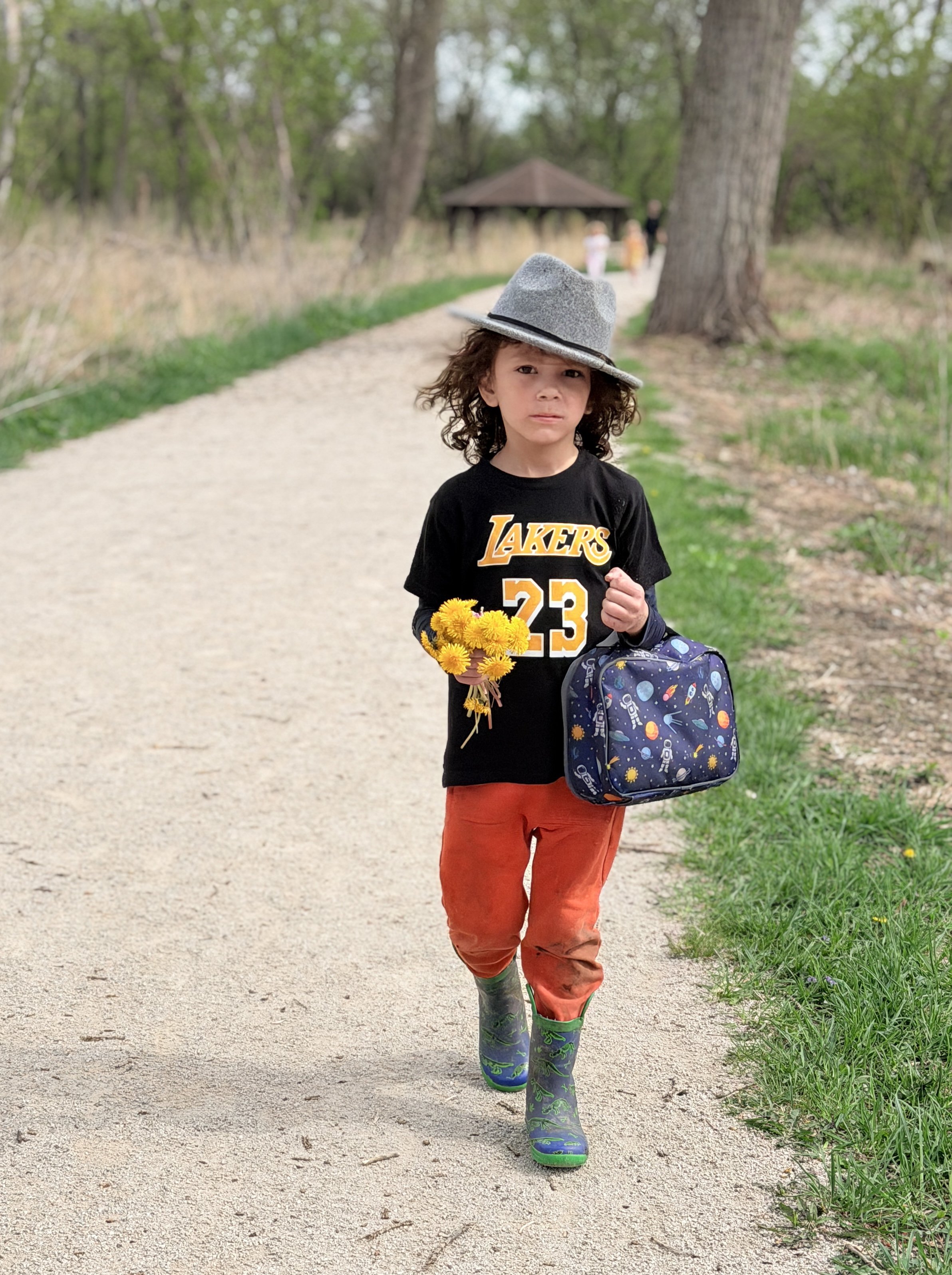 A young boy with curly hair wearing a grey hat, a black Lakers jersey, red pants, and colorful rubber boots, walking on a gravel path in a park holding yellow flowers and a blue backpack.