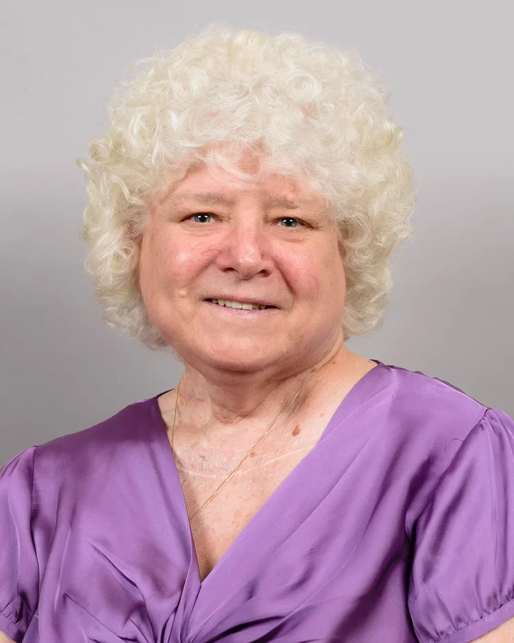 Portrait of an elderly woman with curly white hair, wearing a purple dress, smiling subtly against a plain gray background.
