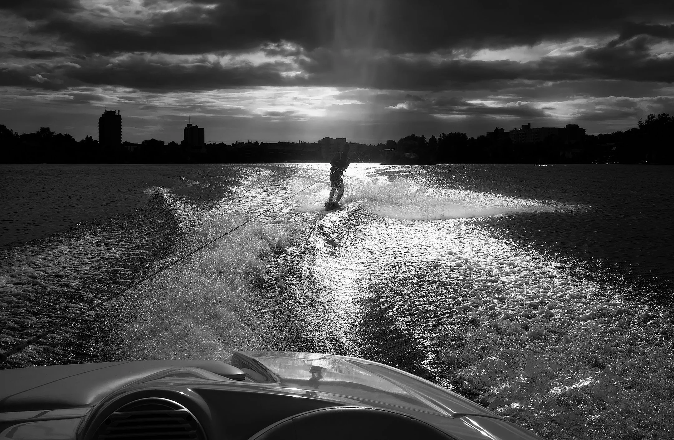 A person wakeboarding on a river, viewed from inside a boat. The sky is cloudy with the sun partially visible, and the city skyline is in the distance.
