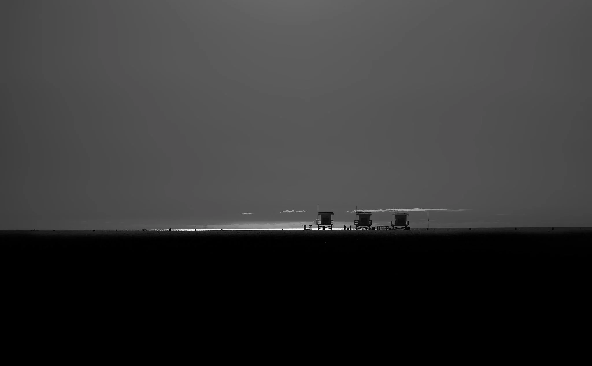 Black and white photo of three lifeguard towers on a beach with a calm ocean and a sky with a few clouds in the background.