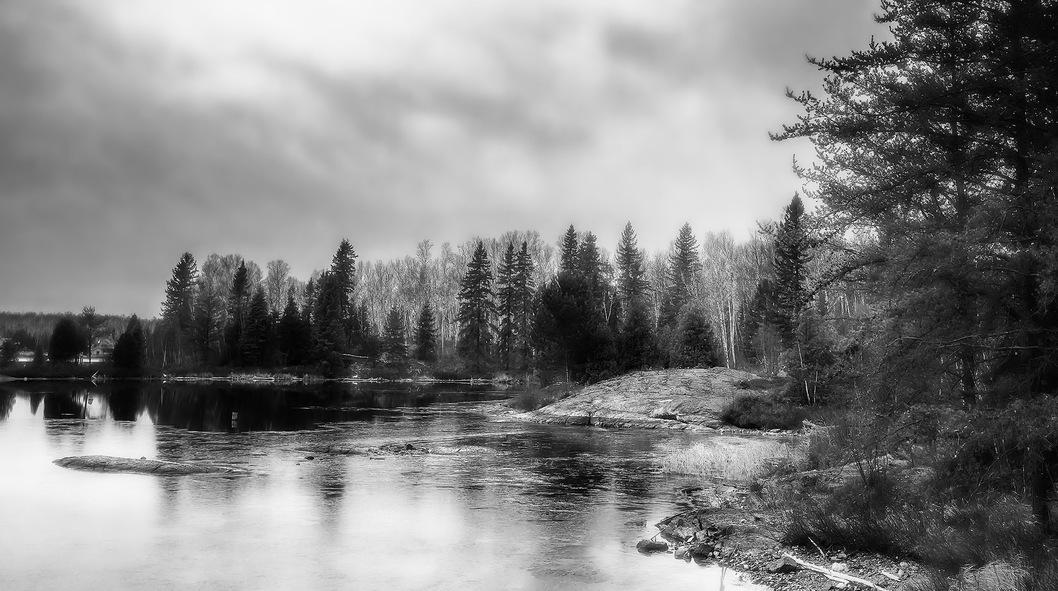 Black and white photograph of a tranquil river flowing through a forested area with dense trees and rocky shoreline under a cloudy sky.