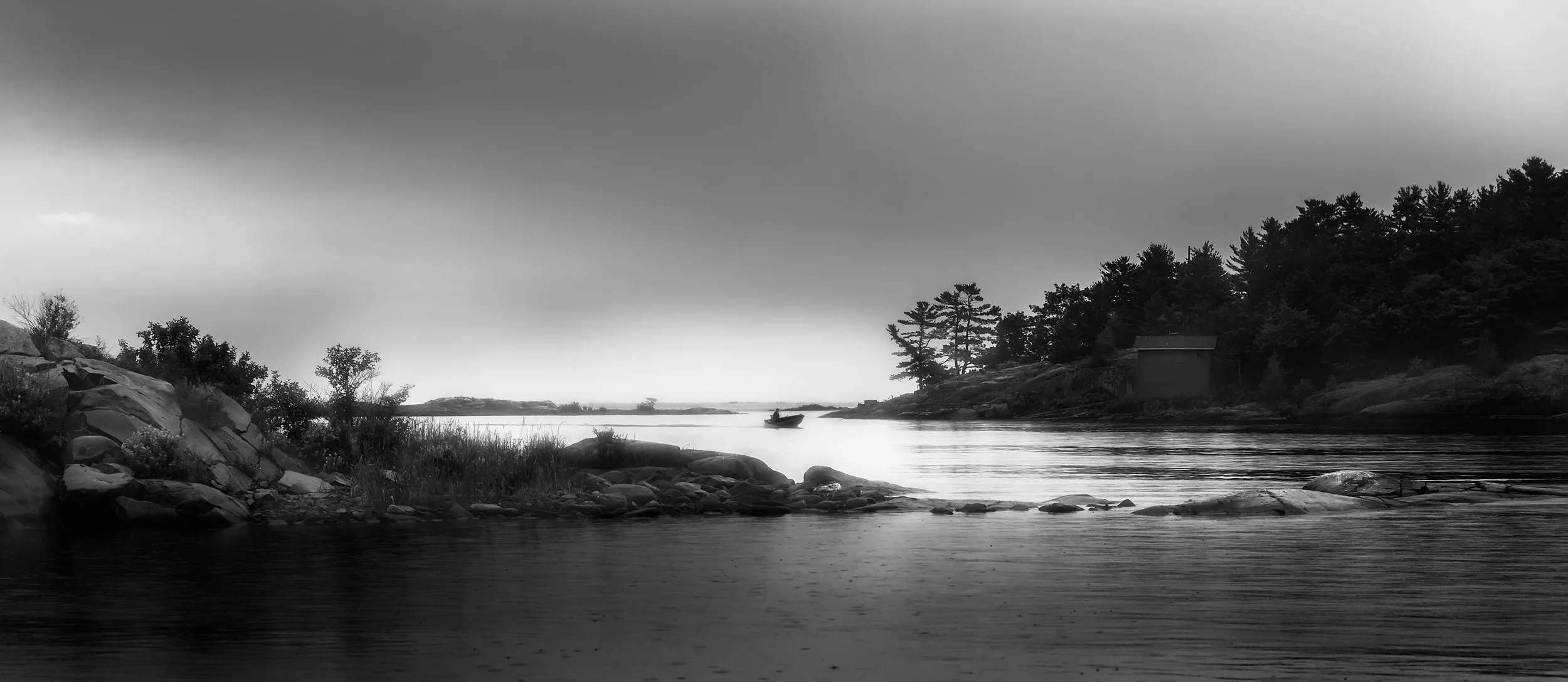 A black and white photo of a river with a small boat and a dense forest on the right side, rocky shoreline on the left, and a cloudy sky.