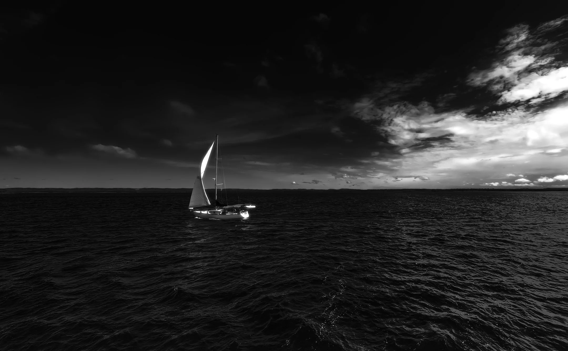 A sailboat with a single sail sailing on dark water under a cloudy sky.