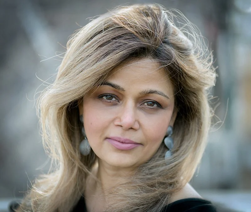 A woman with blonde, wavy hair and earrings looking at the camera outdoors.