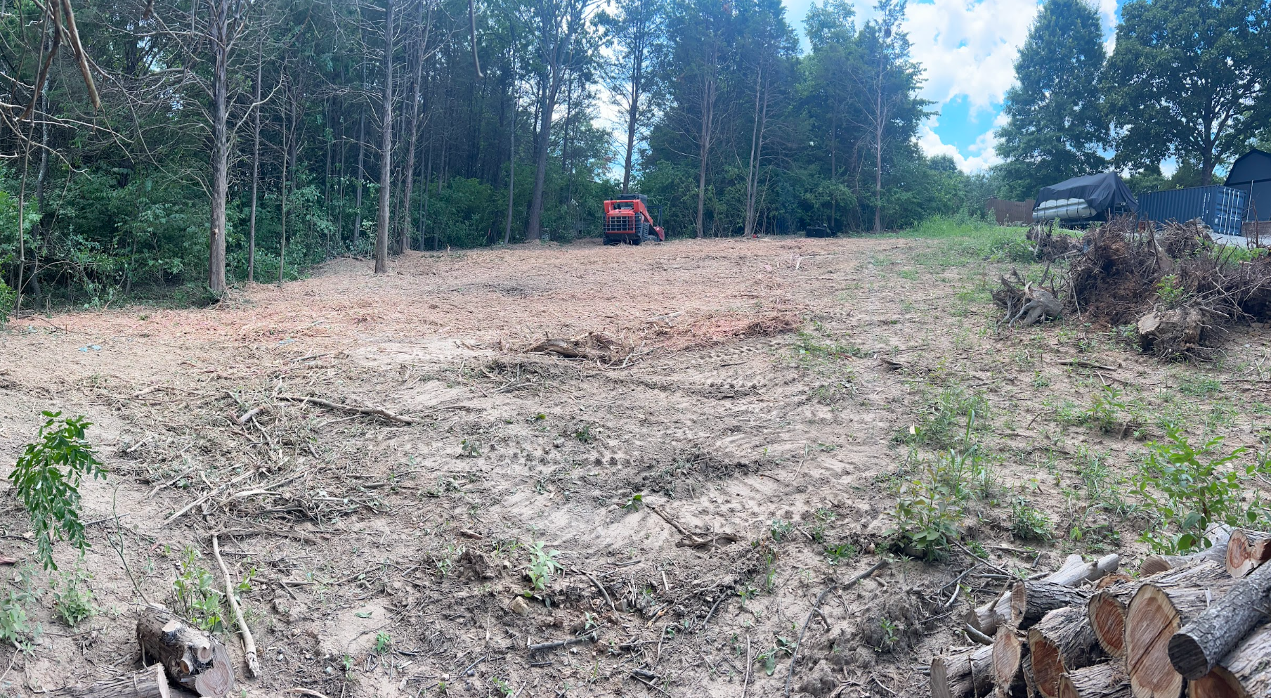 Cleared plot of land with scattered branches, wood logs in the foreground, trees, and a small excavator in the background, under a partly cloudy sky.