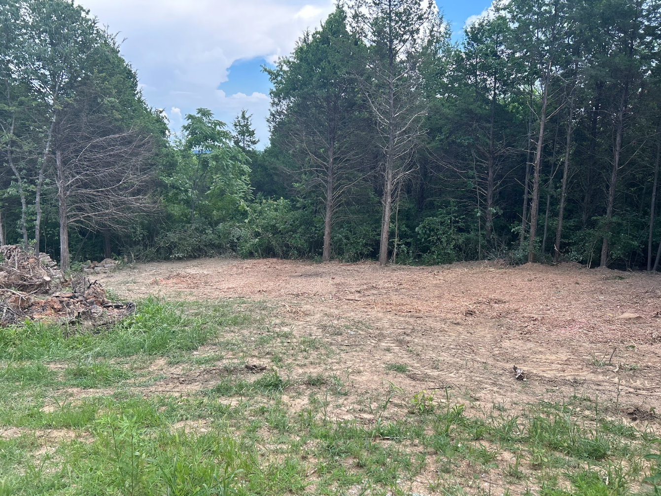 Cleared land with patches of grass, dirt, and trees in the background under a partly cloudy sky.