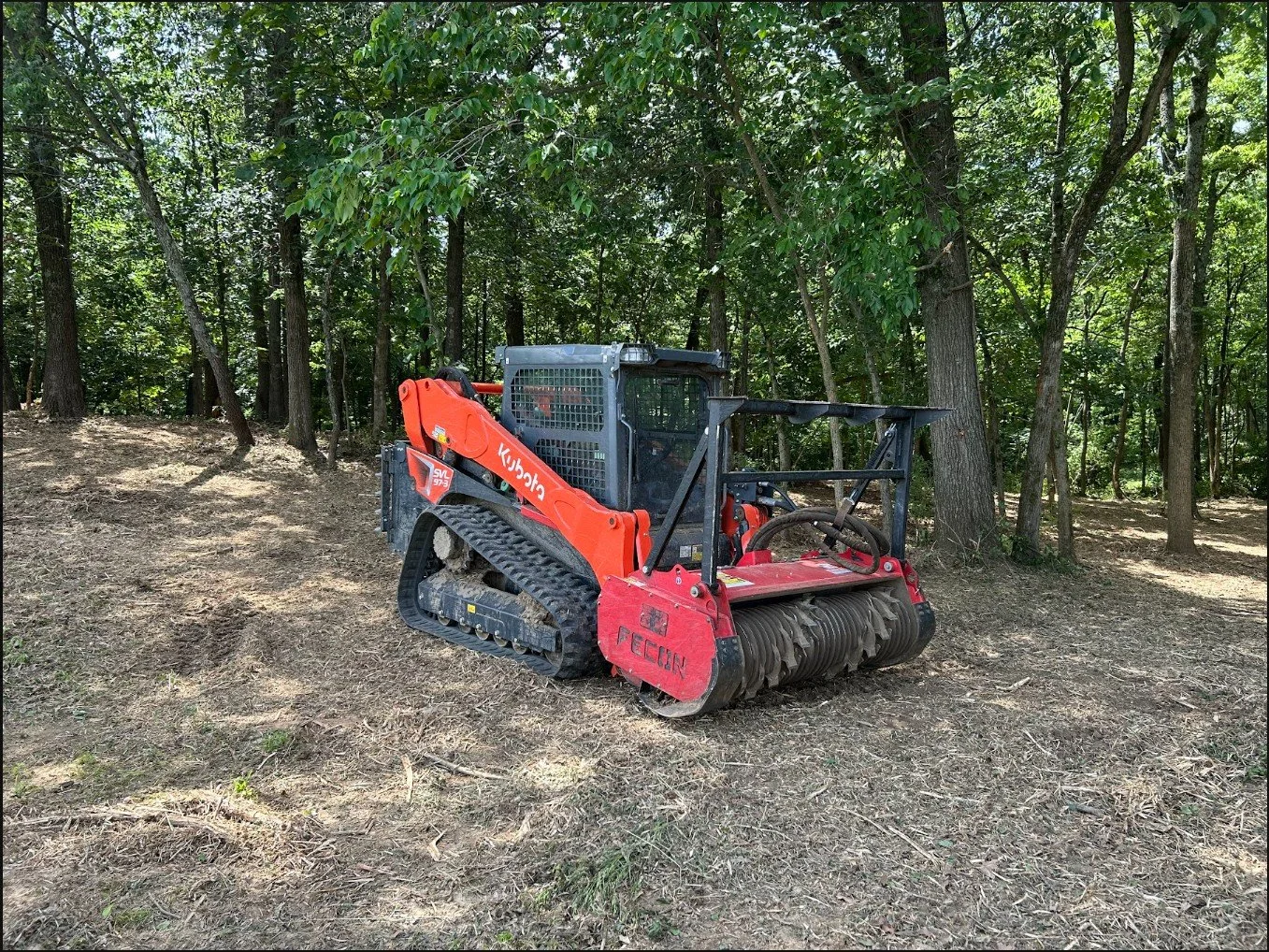 Red Kubota tracked mulcher machine in a wooded area with trees and dirt ground.