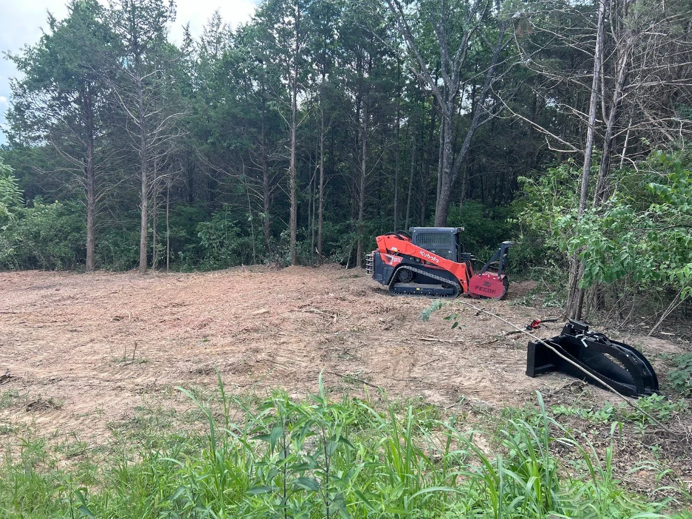 A small orange and black bulldozer with tracks, parked on a cleared patch of land near a wooded area, with green grass in the foreground.