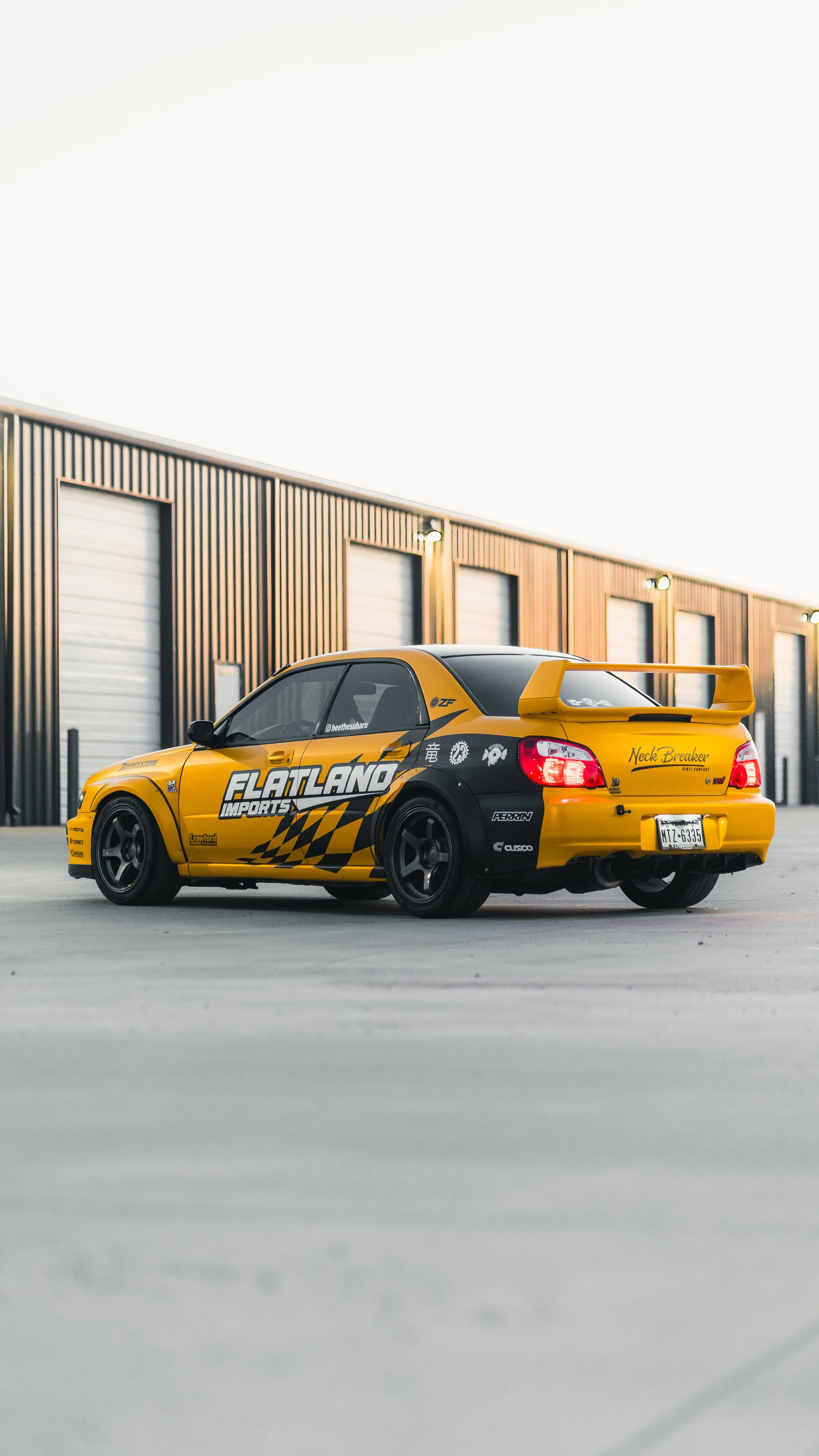 Yellow and black modified sports car with decals and spoilers parked in front of a industrial building with large garage doors.