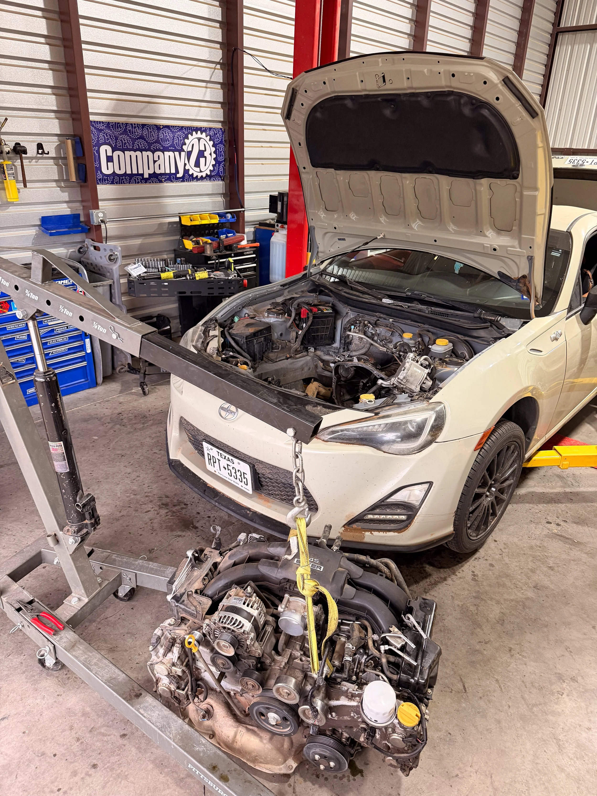 A car with its hood open in a garage, with an engine removed and placed on a metal cart. The garage has various tools and a banner that reads 'Company 23' on the wall.