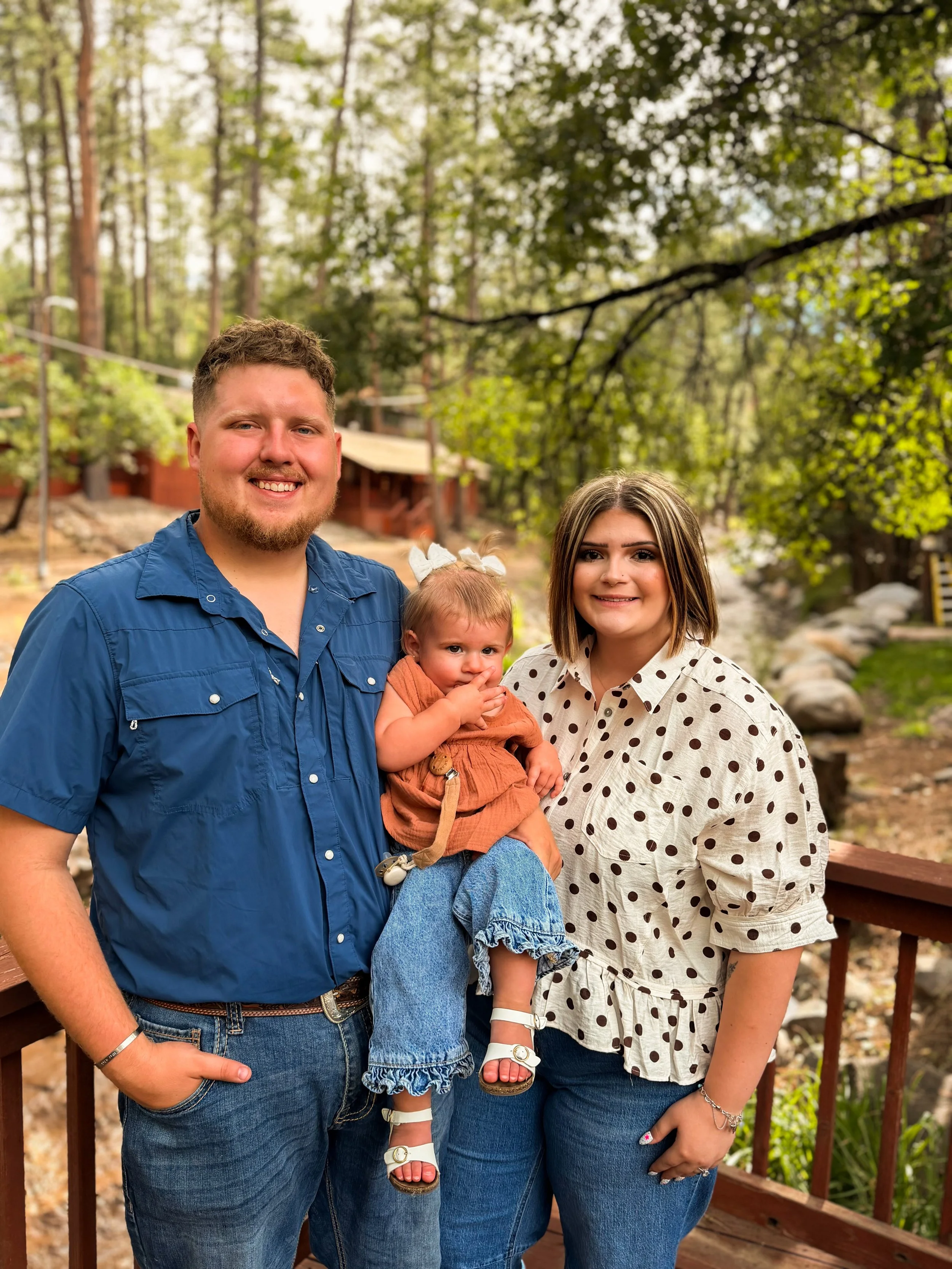 A smiling family of four standing outdoors on a wooden deck in a forested area with tall trees and green foliage. The father is wearing a blue shirt, the mother is wearing a beige polka dot shirt, and the two children are being held, one of whom has a bow in her hair and is wearing a brown dress with jeans.