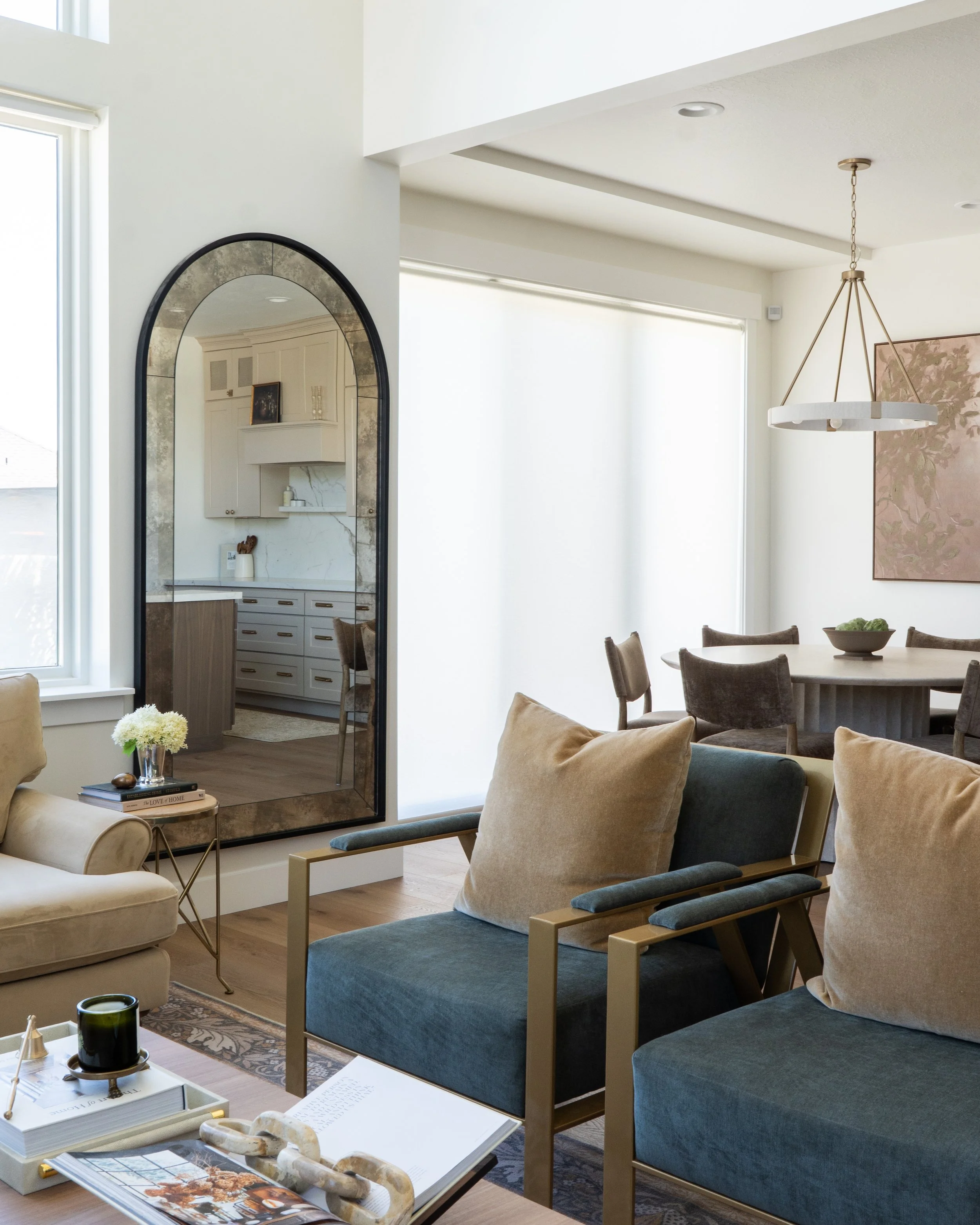 Living room with beige and blue armchairs, a round dining table with chairs, a modern chandelier, a large window with white blinds, and a mirror reflecting a kitchen area with white cabinetry and a marble countertop.