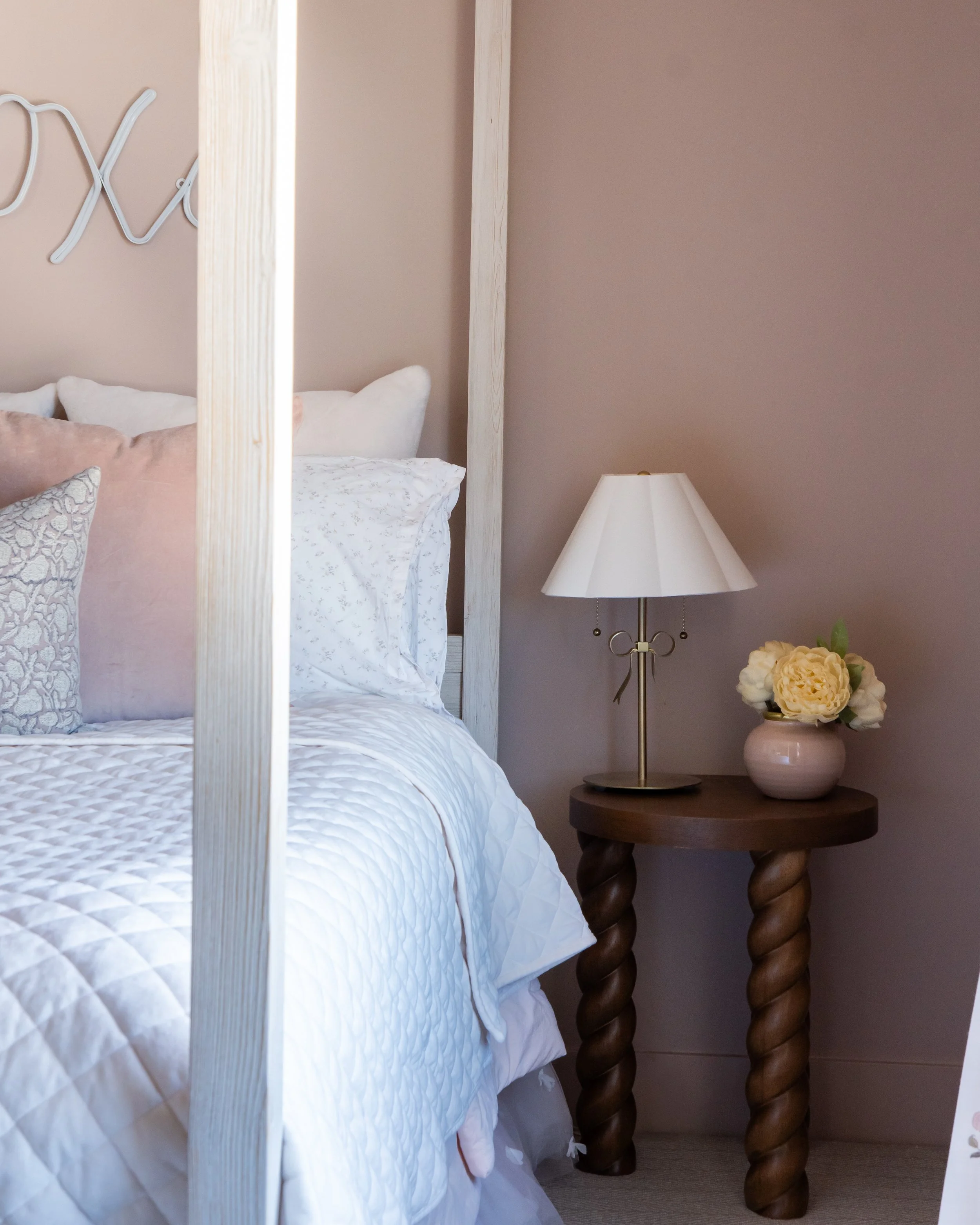 A close-up of a bedroom with a bed, a beige pillow, and a round wooden side table with a white table lamp and a beige vase with white flowers.
