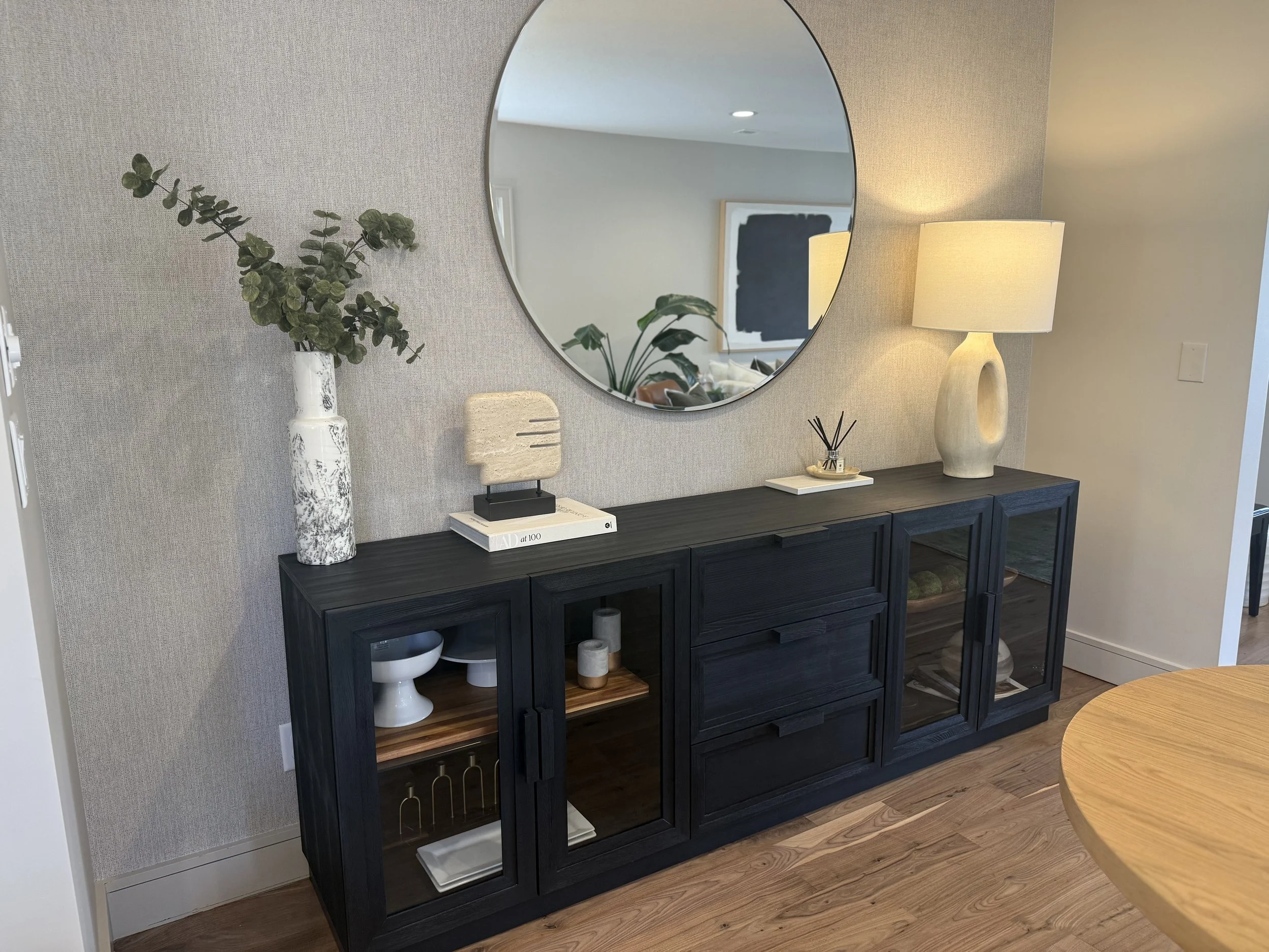 A black wooden sideboard with open shelves and glass cabinet doors, topped with potted greenery, decorative objects, and a large round mirror reflecting a part of the room, with a table lamp on the right side.