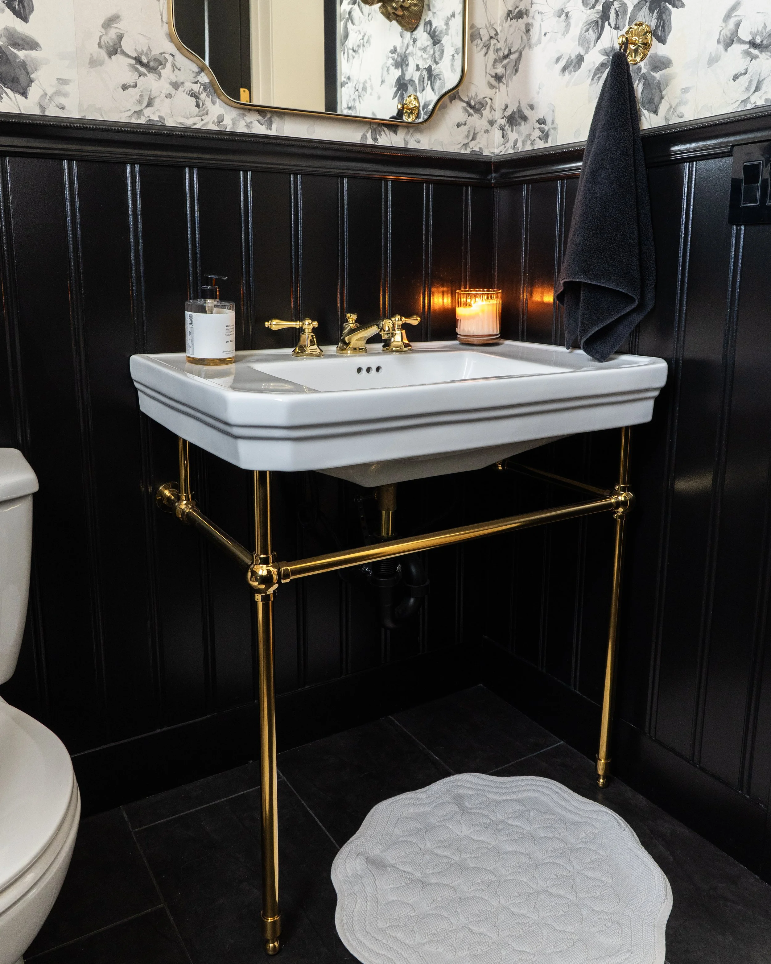 A bathroom sink with gold fixtures and black wainscoting. There is a lit candle and a hand soap dispenser on the sink. A black towel hangs on a gold hook on the wall, above a white textured rug on the black floor.