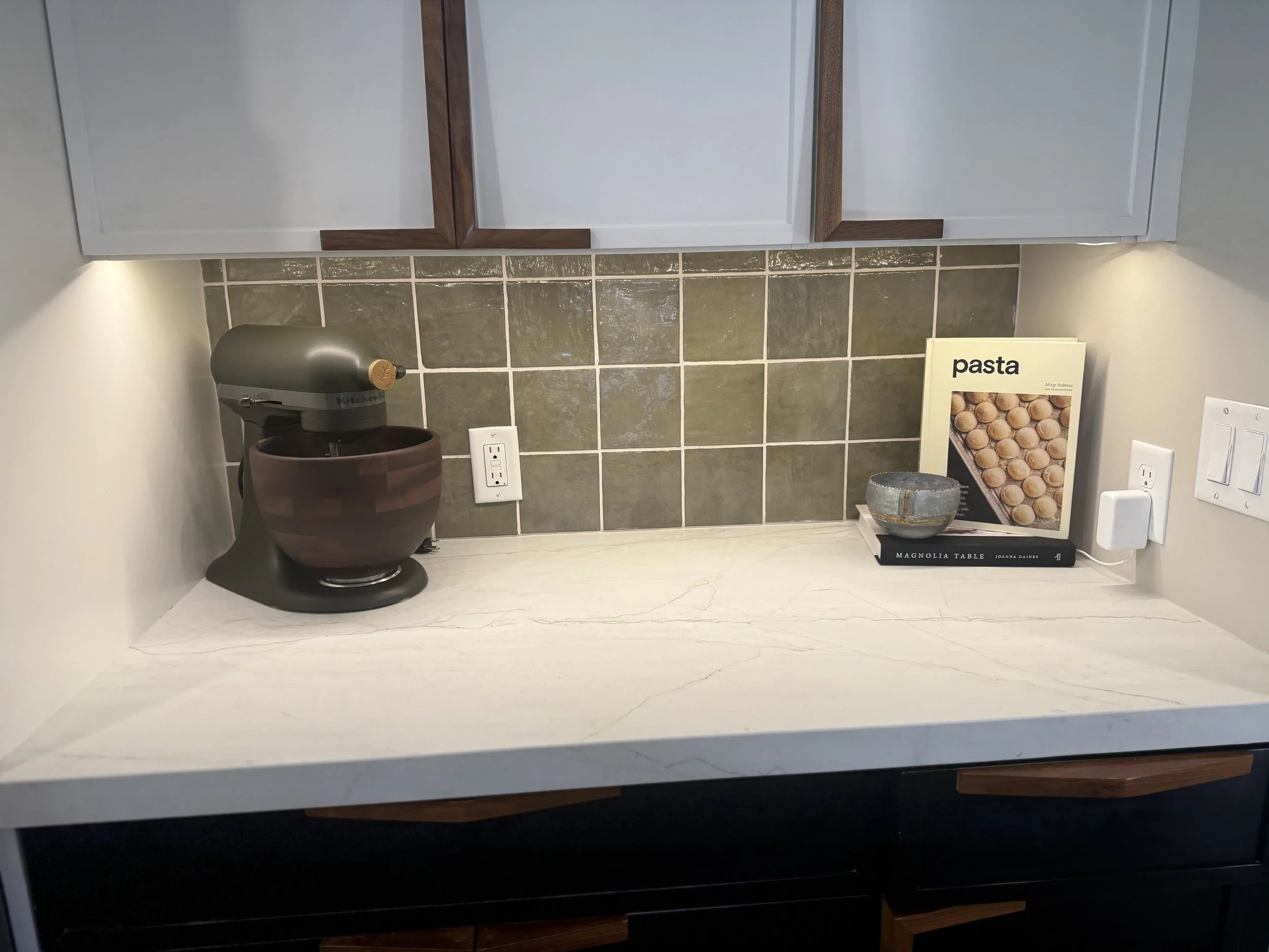 Kitchen countertop with a silver KitchenAid mixer, a brown ceramic bowl, a gray ceramic bowl, a stack of books titled 'Pasta' and 'Magnolia Table'. There is a leafy tile backsplash, an electrical outlet, and a white wall with a light switch.