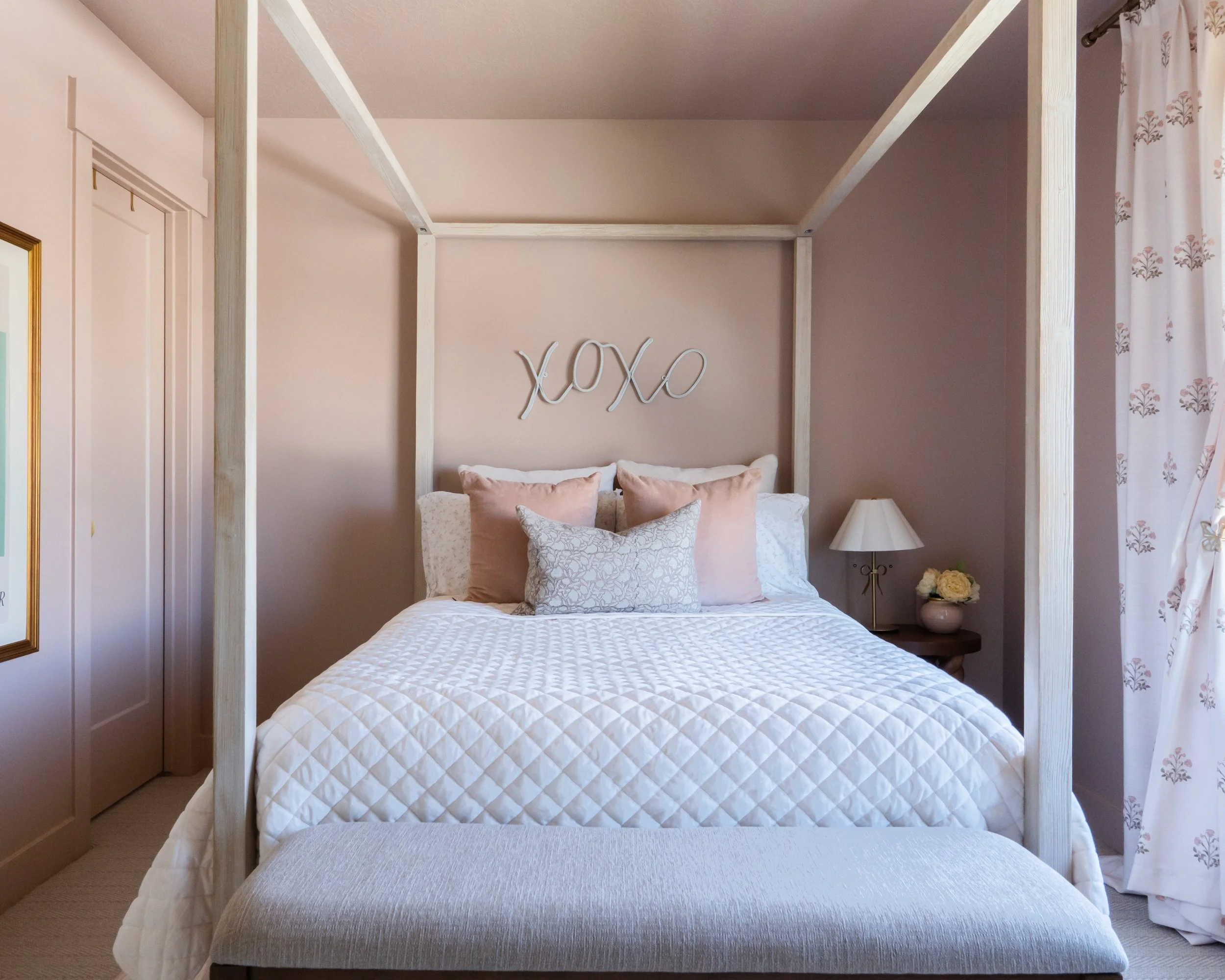 A bedroom with a four-poster bed, decorative pillows, a bedside lamp, and curtains with floral patterns.